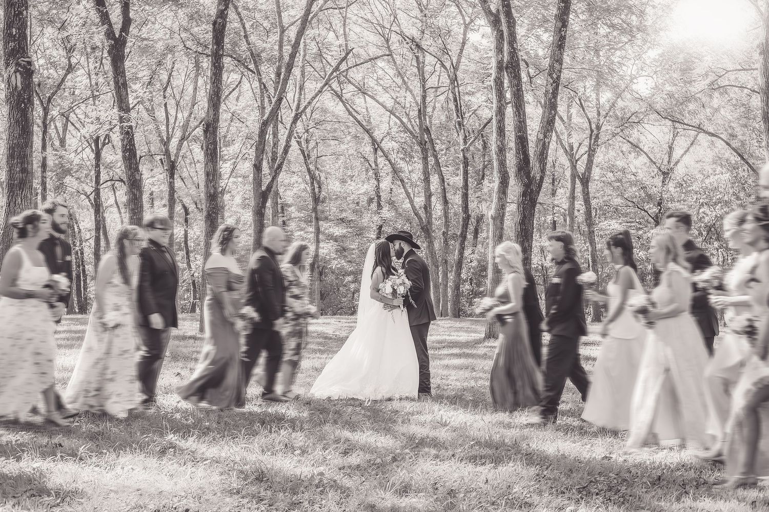 Black and white photo of a bride and groom kissing surrounded by wedding party in a wooded outdoor setting.