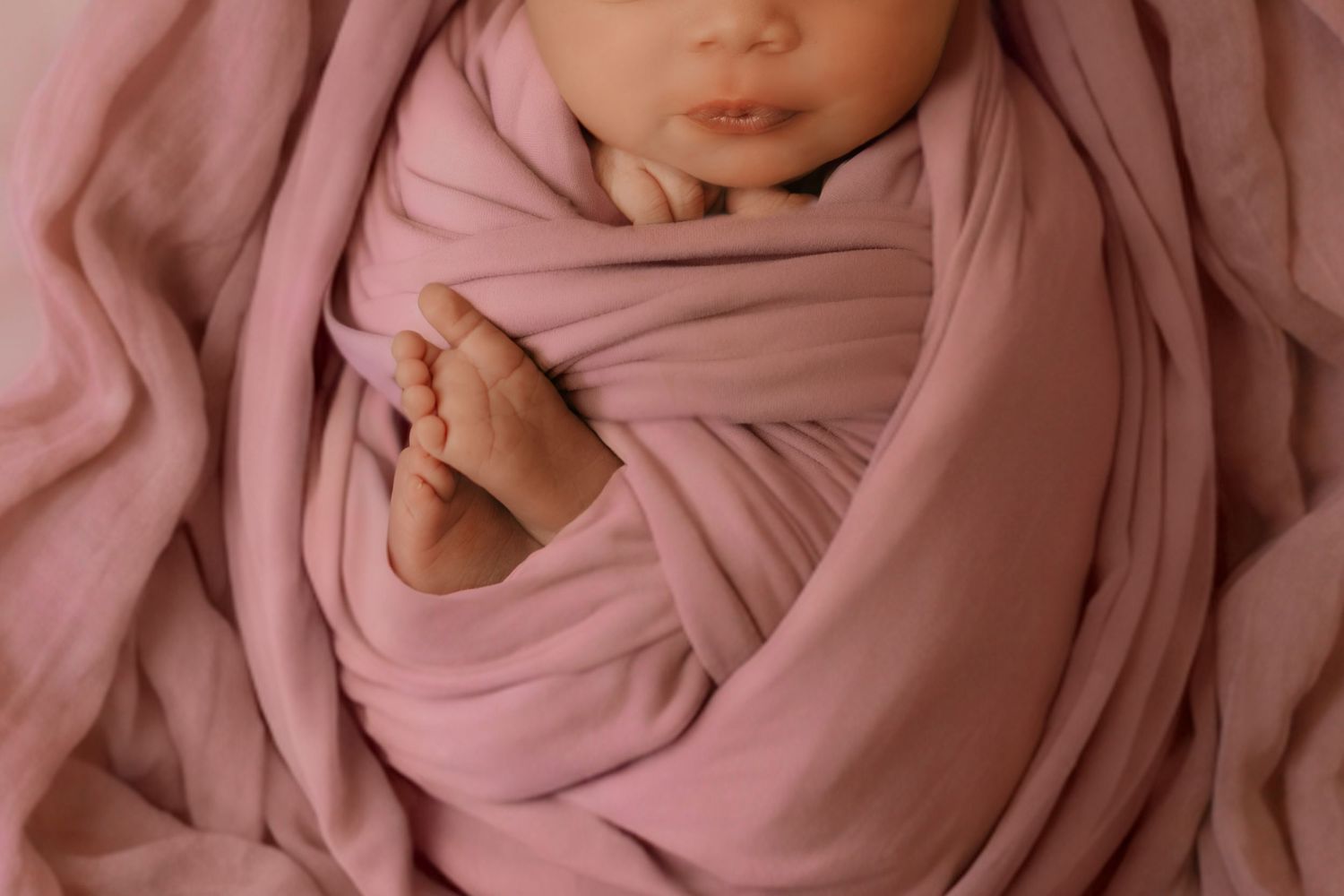 A close-up of a swaddled newborn baby in a pink wrap with tiny hands resting gently on their chin.