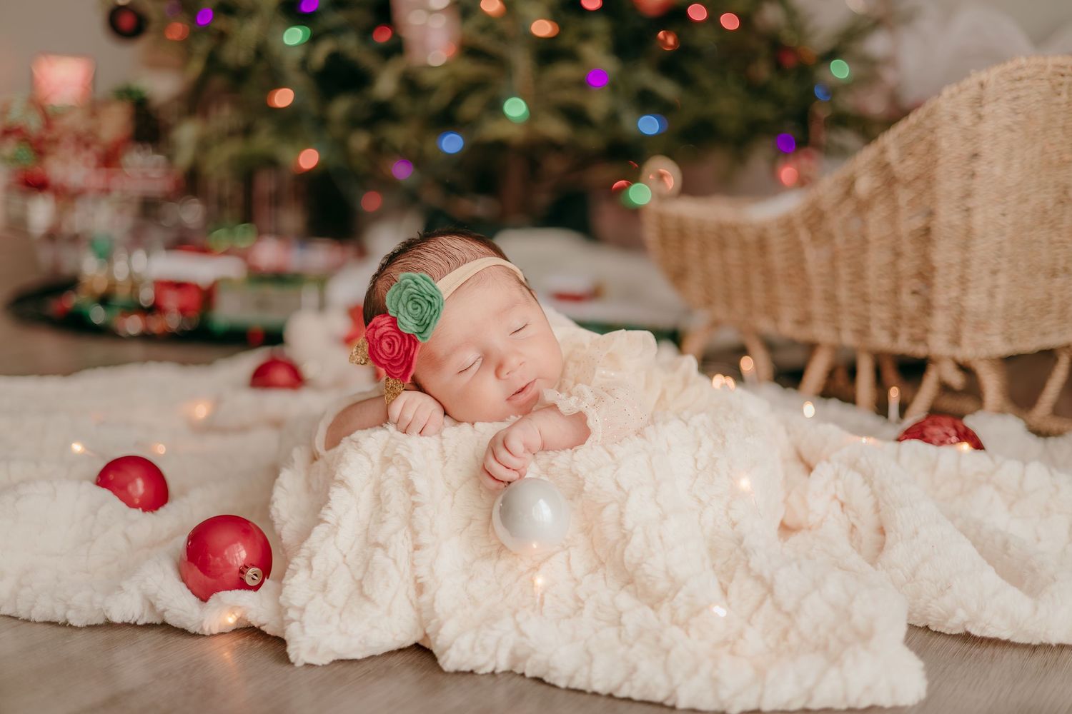 Sleeping newborn baby swaddled in cream blanket surrounded by Christmas ornaments near a lit tree.