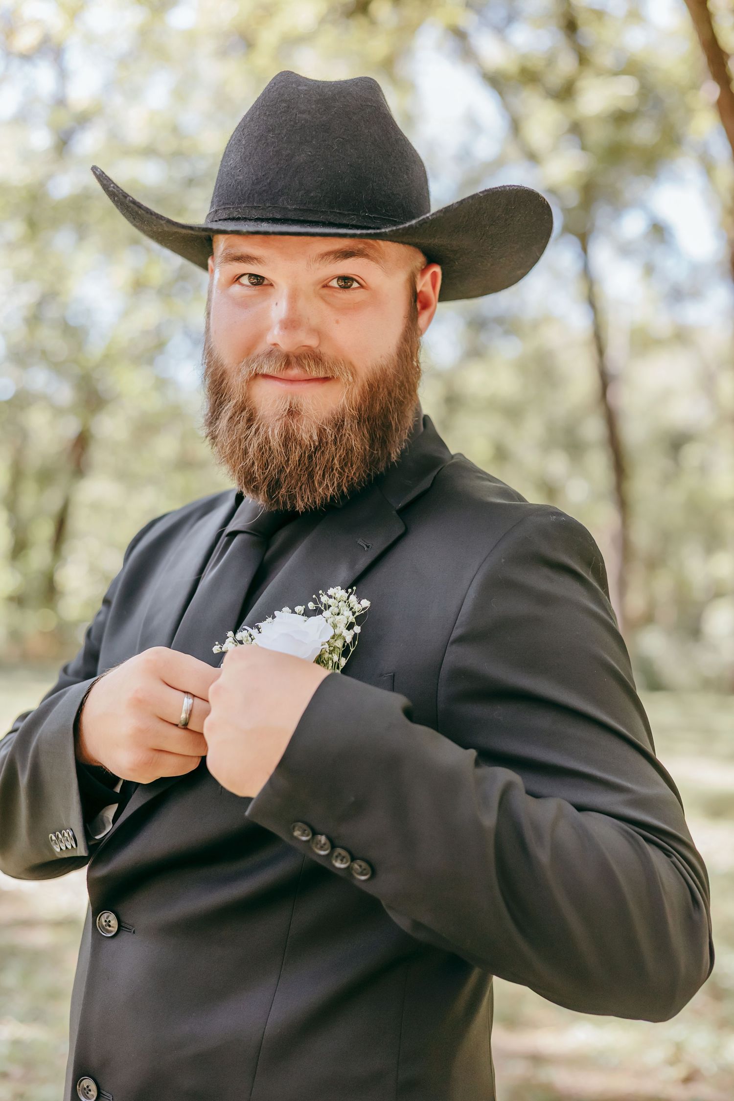 Bearded groom in black cowboy hat and suit adjusting jacket with floral boutonniere in forest.