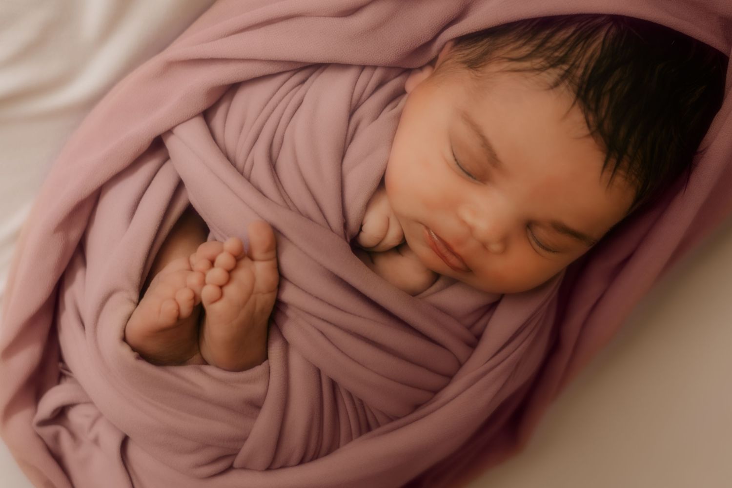 Close-up of a sleeping newborn baby wrapped in a mauve pink swaddle with tiny hands visible.