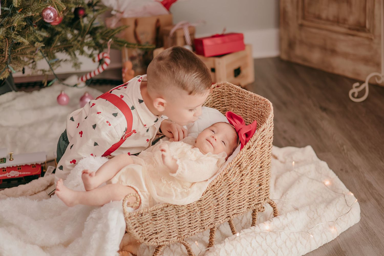 Older sibling kissing sleeping newborn baby in wicker bassinet near Christmas tree with gifts.