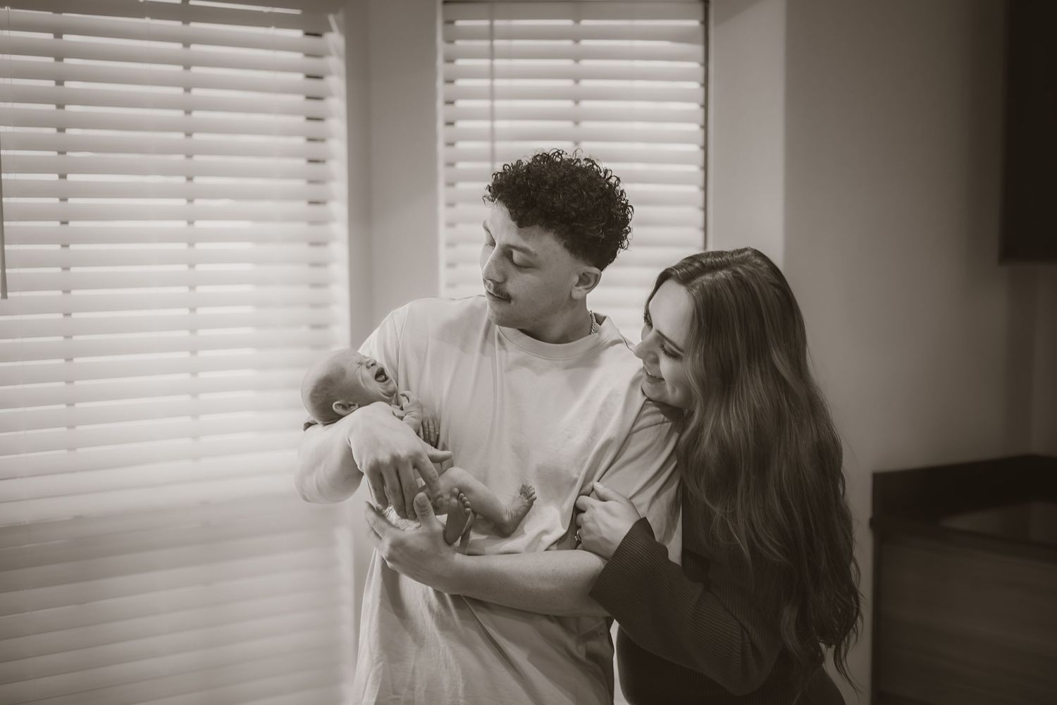 Black and white photo of new parents lovingly gazing at their newborn baby by a window.