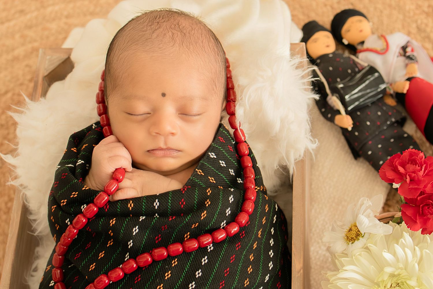 Newborn baby wrapped in black traditional fabric with red beaded necklace sleeping in wooden box with dolls.