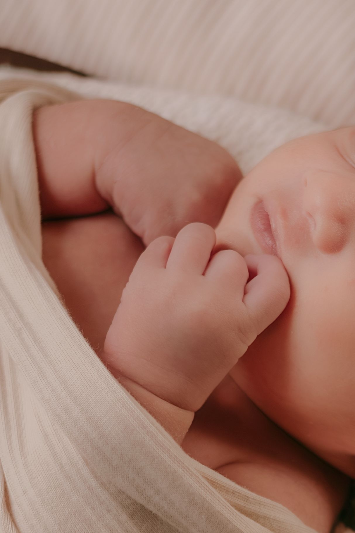 Close-up of sleeping newborn baby tiny hand and lips wrapped in soft cream muslin cloth.