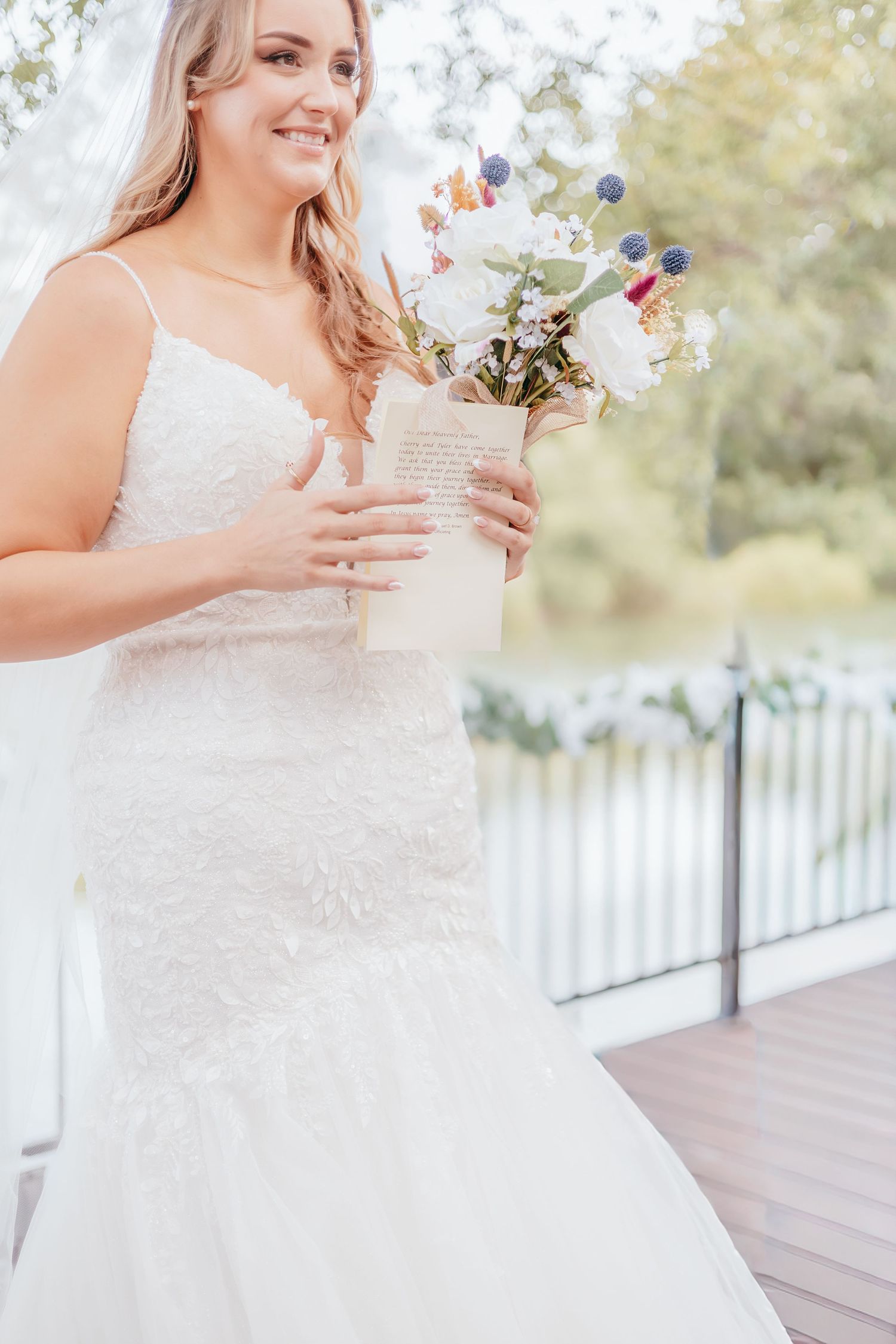 Smiling bride in lace wedding gown holding a floral bouquet with a handwritten note outdoors.