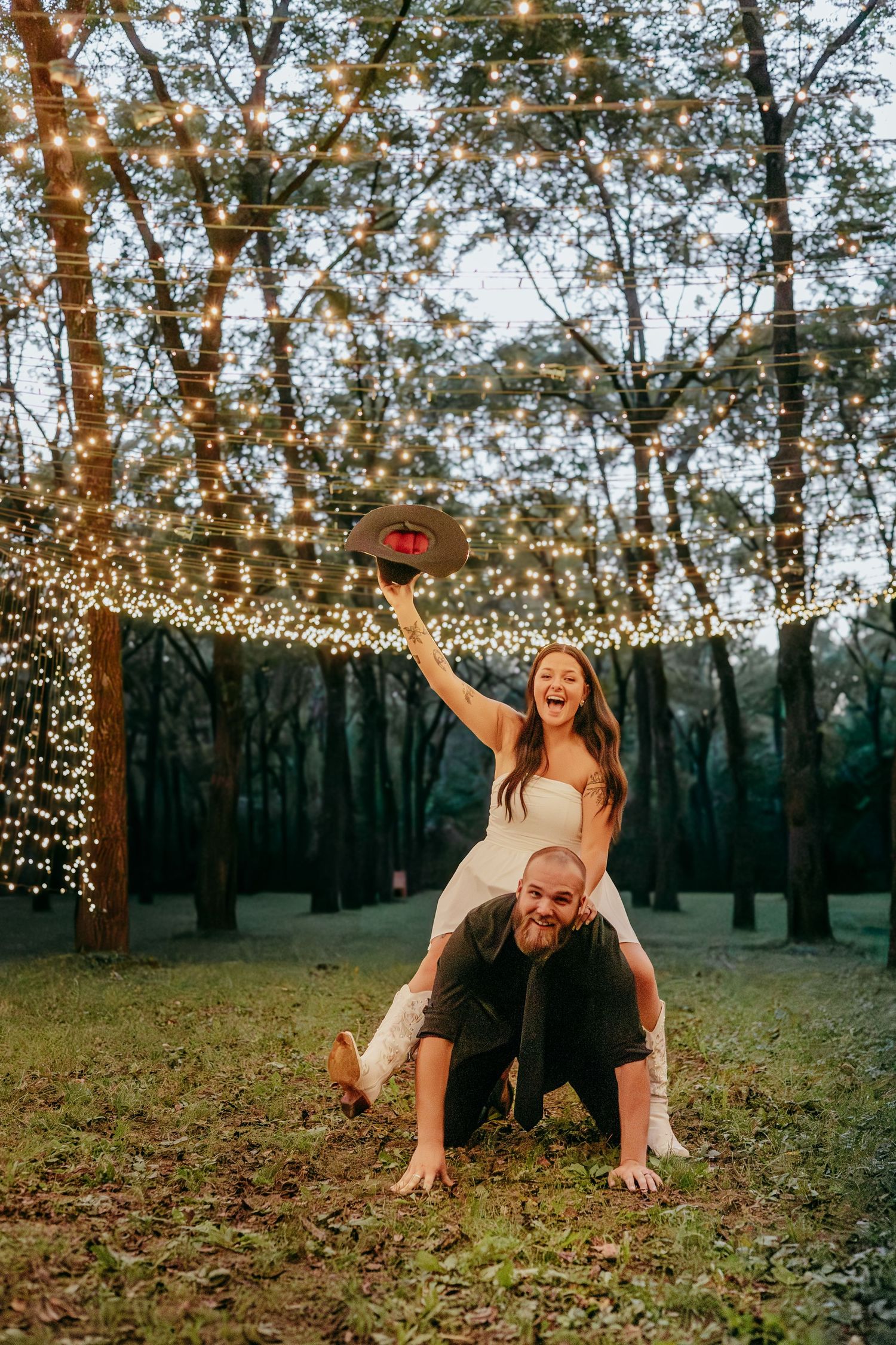 Joyful bride sitting on grooms shoulders holding a cowboy hat under sparkling fairy lights in a forest.