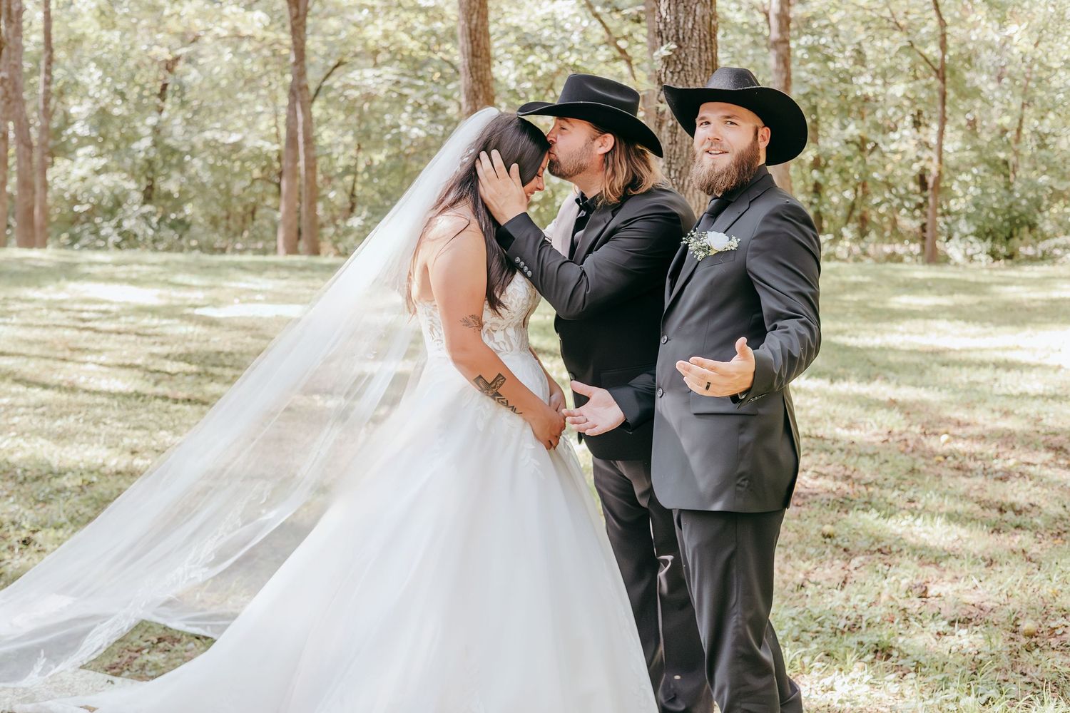 Officiant kisses bride while groom in black cowboy hat reacts humorously during outdoor wedding.