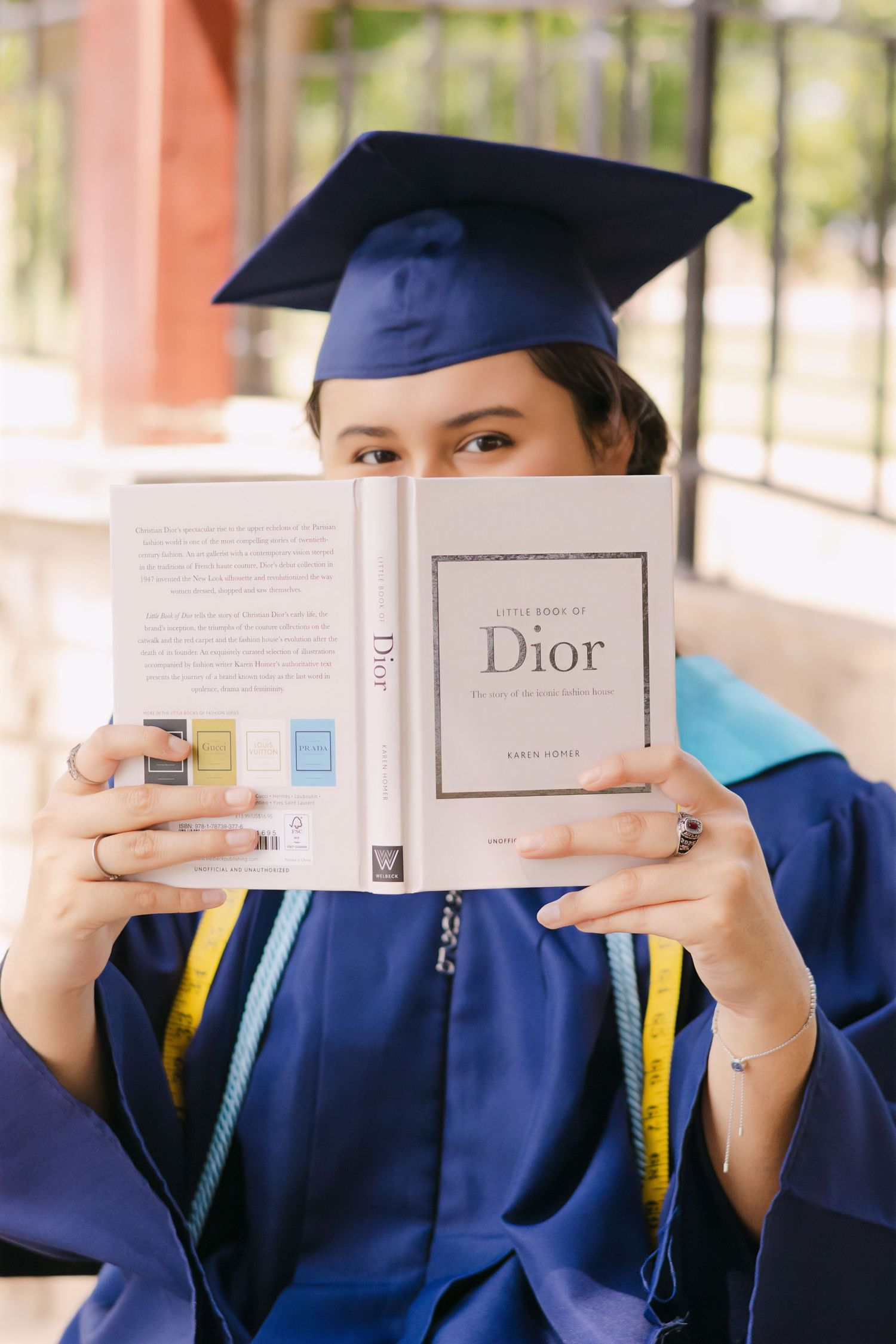 Graduate in blue cap and gown holding Little Book of Dior fashion book over face.