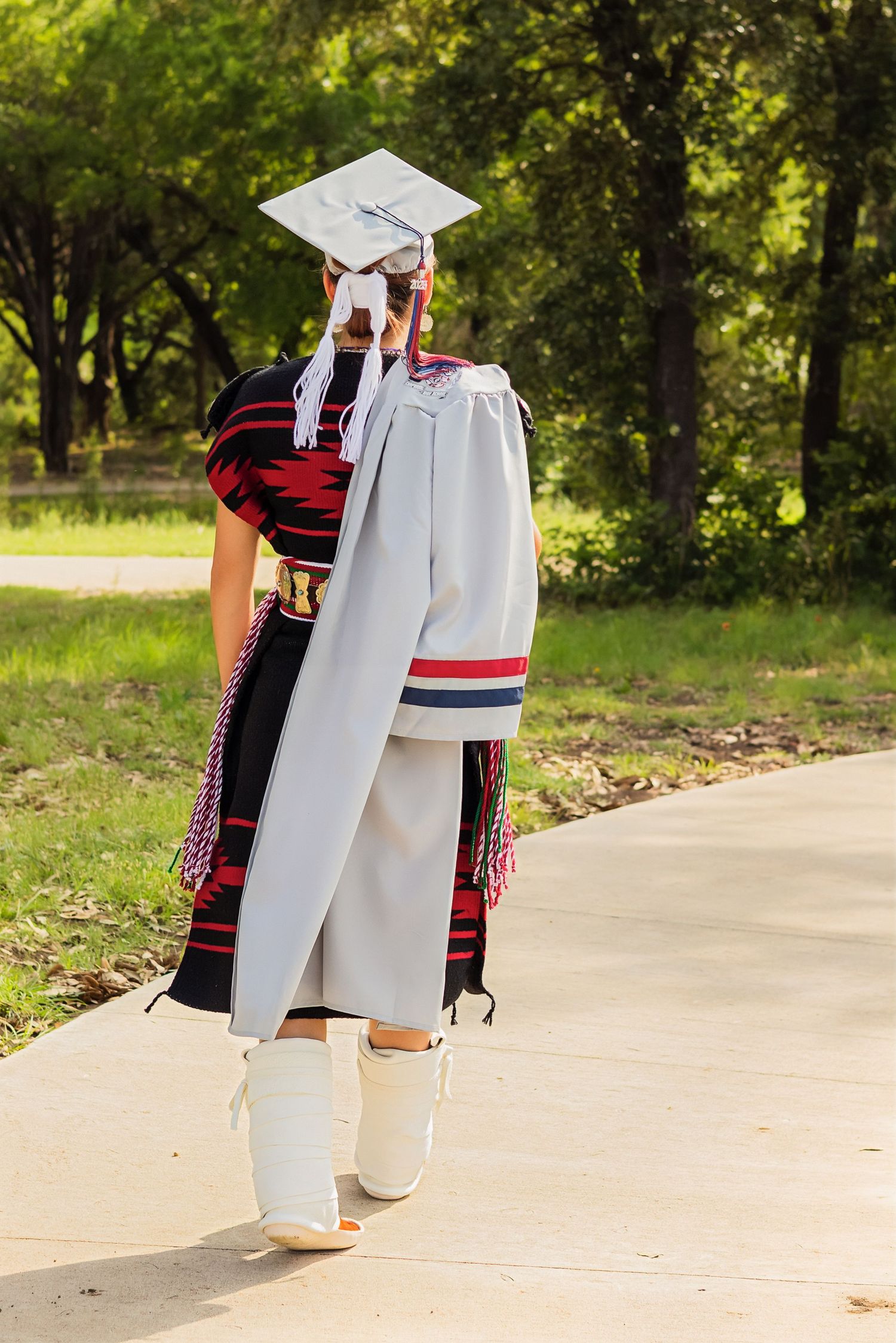 Native American graduate walks away in traditional regalia and white graduation cap on sunny tree-lined path.