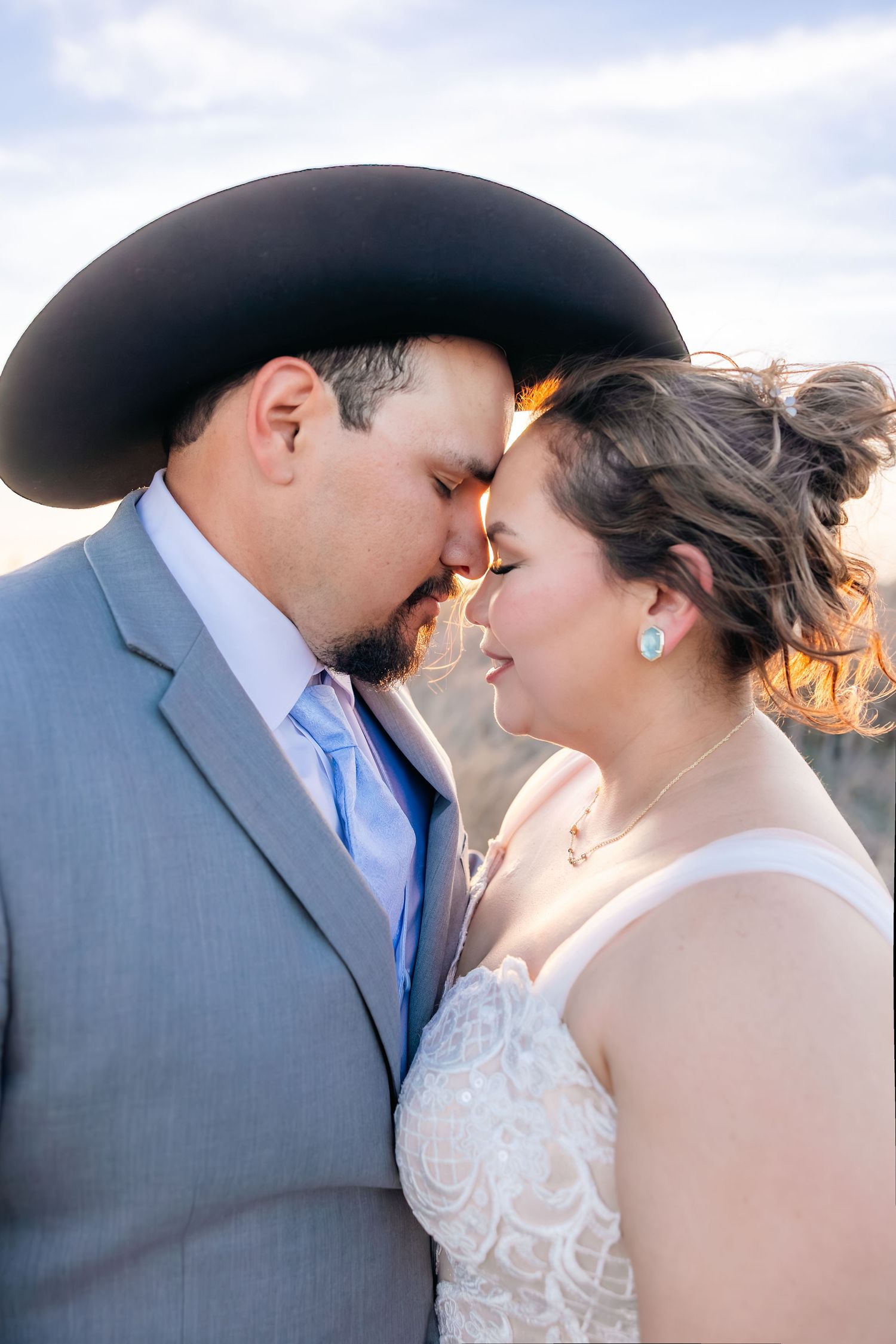 Bride and groom share an intimate nose-to-nose moment, groom in black cowboy hat and gray suit at sunset.