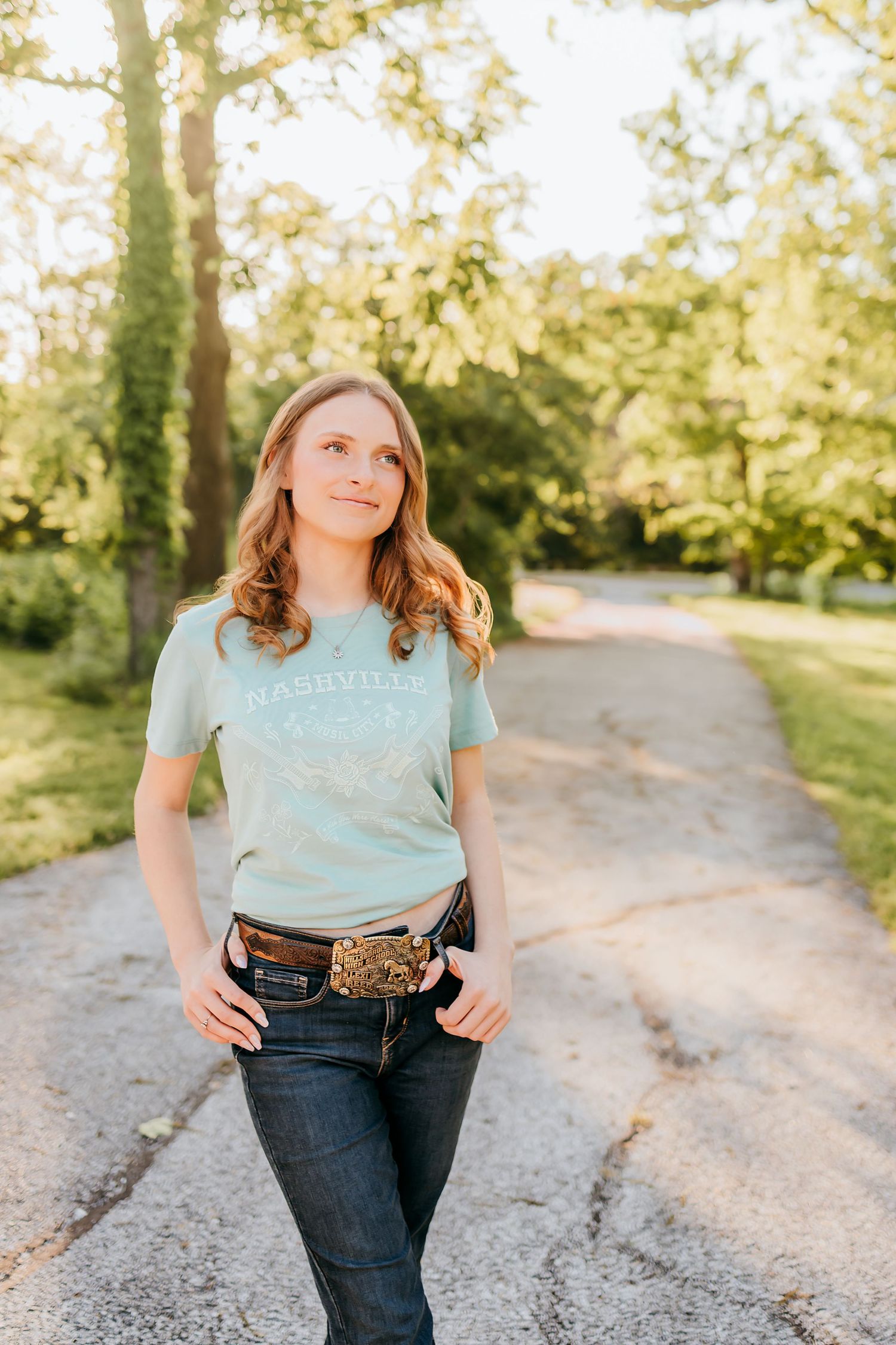 Young woman in Nashville t-shirt and jeans with western belt poses on a tree-lined gravel path in golden sunlight.