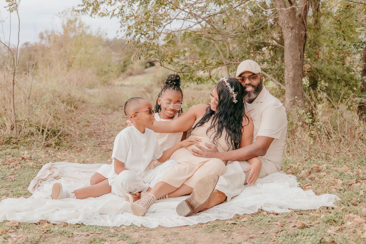 Happy family of four sitting on white blanket outdoors, sharing joyful embrace in natural setting.