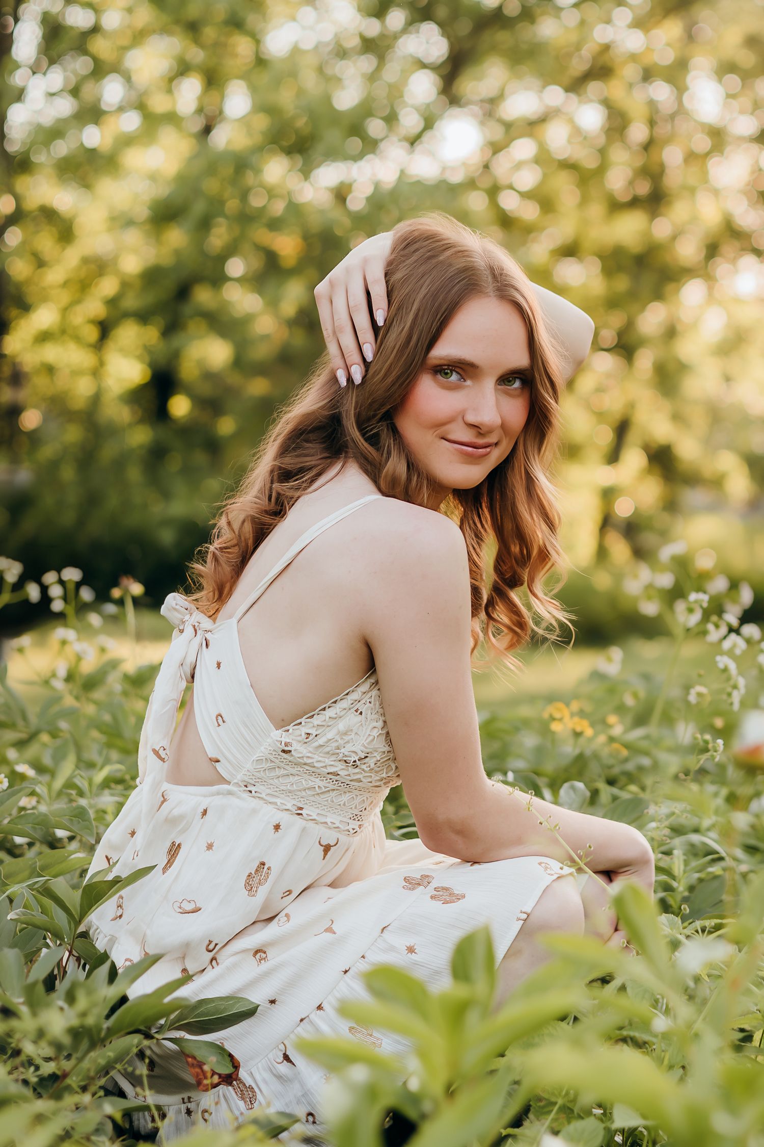 Young woman in floral sundress sitting in green meadow, smiling over shoulder, golden hour sunlight, white headband.