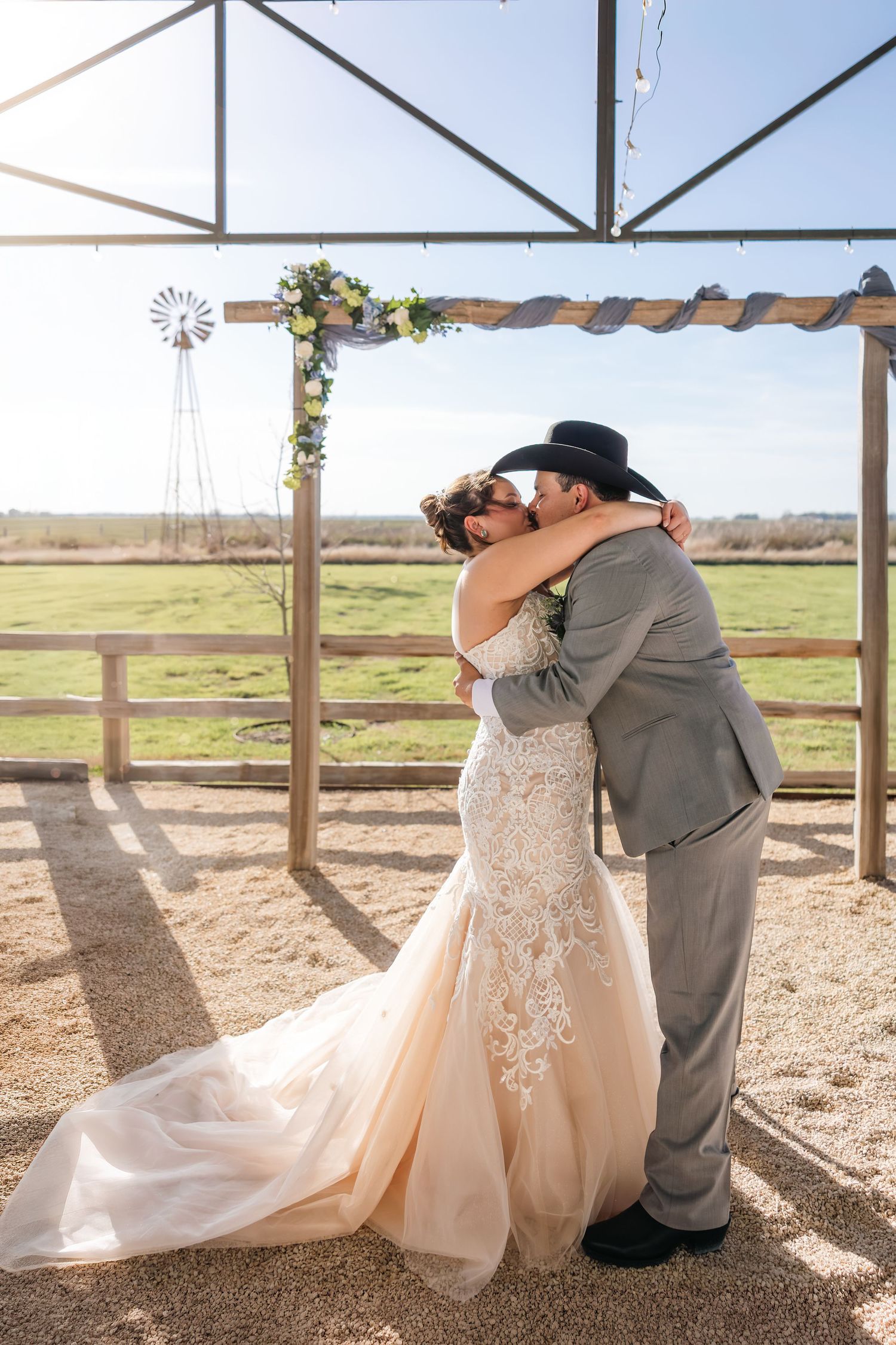 Bride and groom share first kiss under a floral arch at a rustic outdoor farm wedding venue.