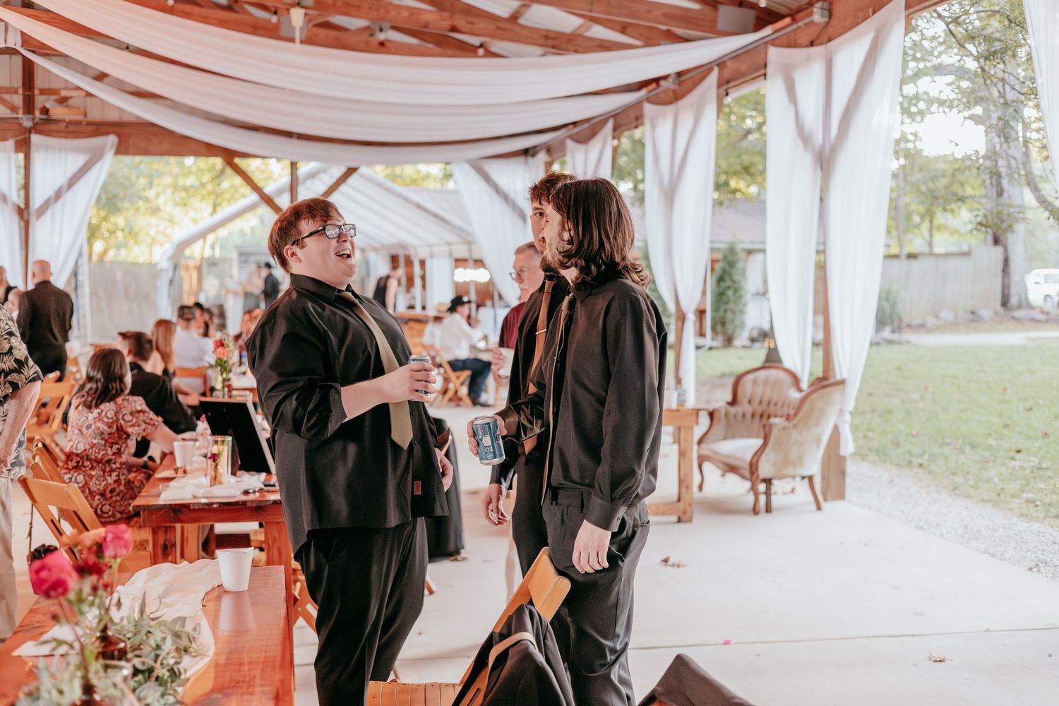 Two guests in black attire chatting under a draped pavilion at a rustic outdoor wedding reception.