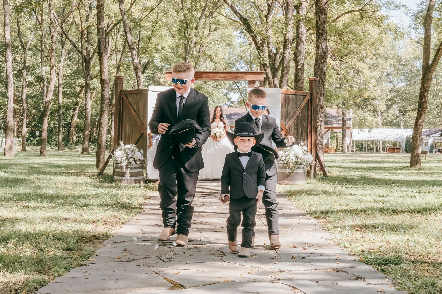 Two men in sunglasses escort young ring bearer in black suit down outdoor wedding aisle.