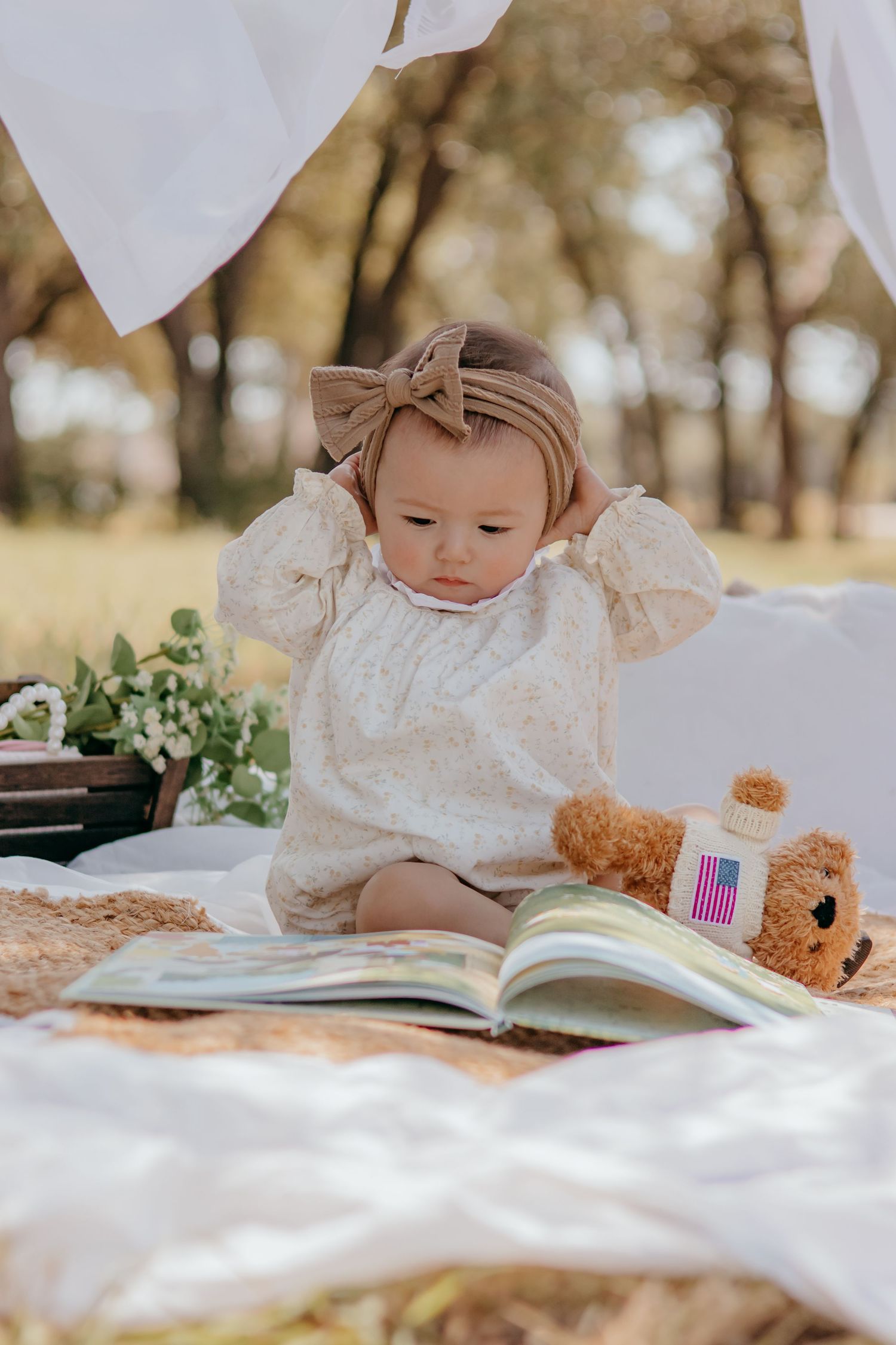 Adorable baby girl in floral romper and bow headband reading a book outdoors with a teddy bear on a white blanket.