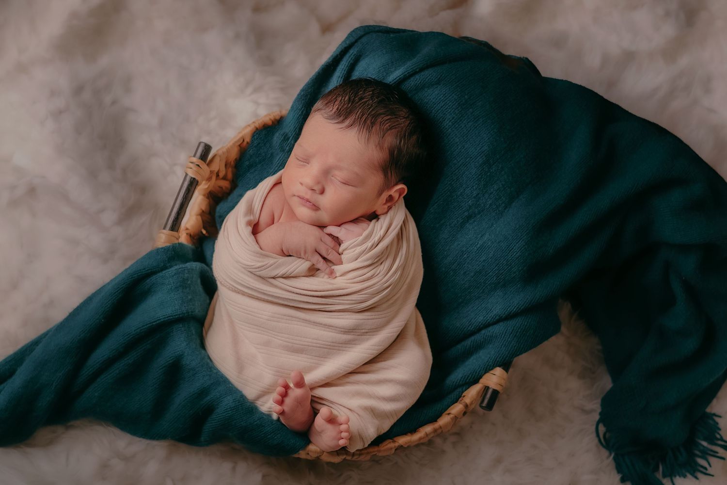 Sleeping newborn baby swaddled in cream wrap, nestled in wicker basket with teal blanket.
