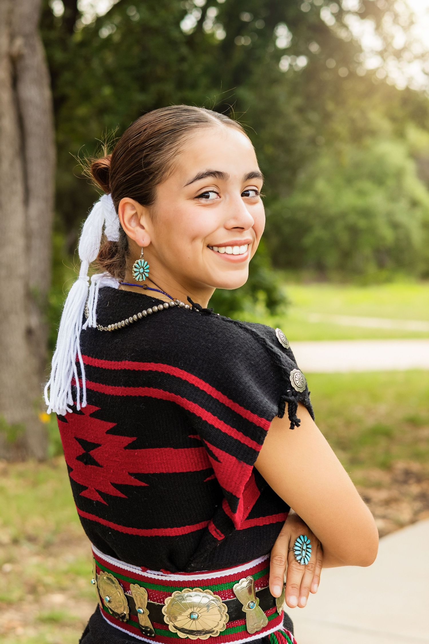 Young woman smiling in traditional Native American attire with turquoise jewelry and geometric patterned outfit outdoors.