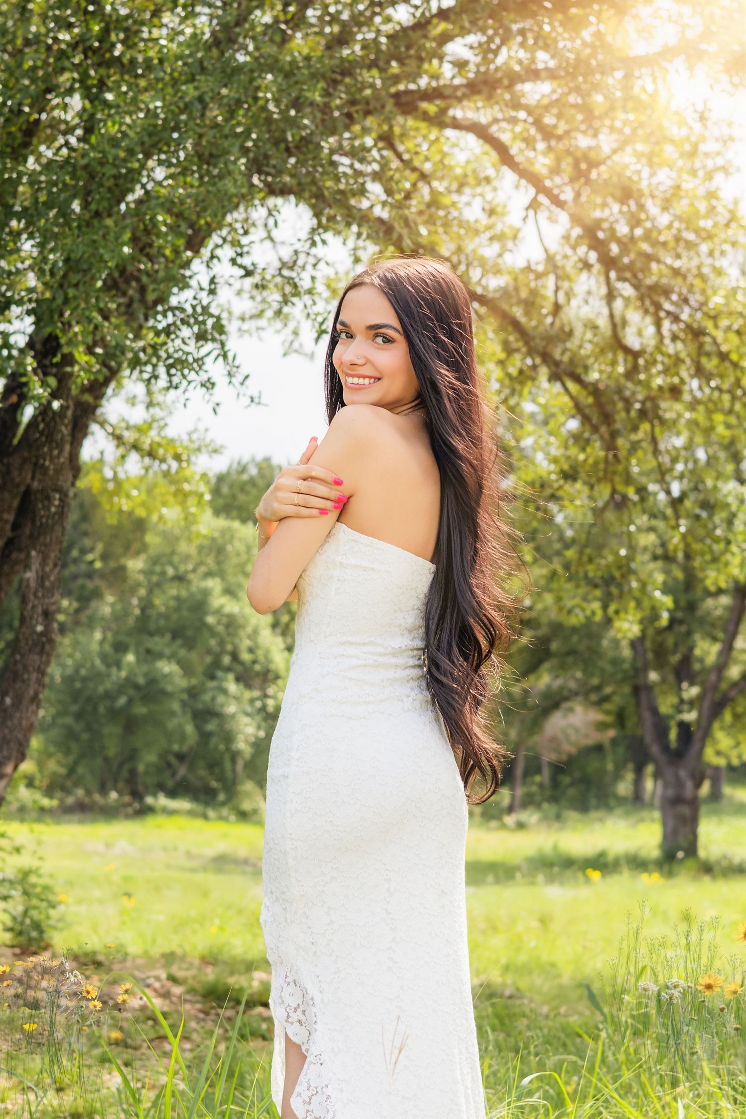 A young woman in a white strapless lace gown smiles over her shoulder outdoors among green trees in sunlight.