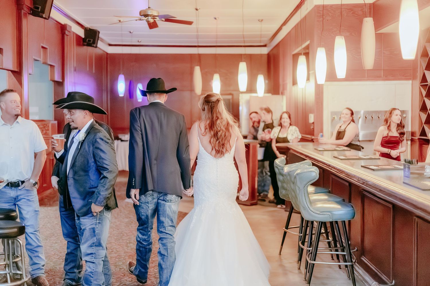 Bride and groom in western attire walk hand-in-hand through a rustic bar venue during their wedding reception.