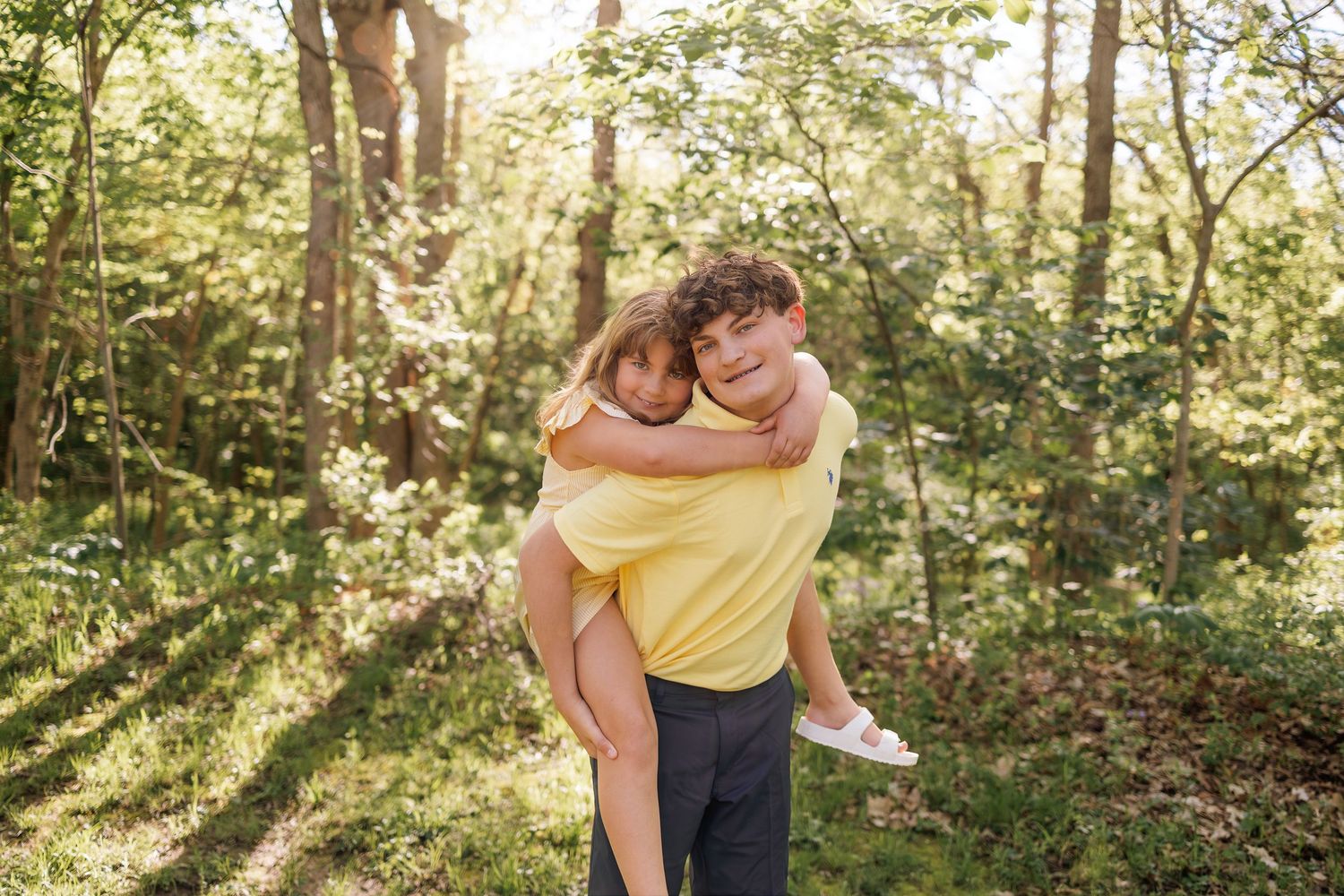 Young couple embracing in a sunny forest, man in yellow shirt carrying woman piggyback among green trees.