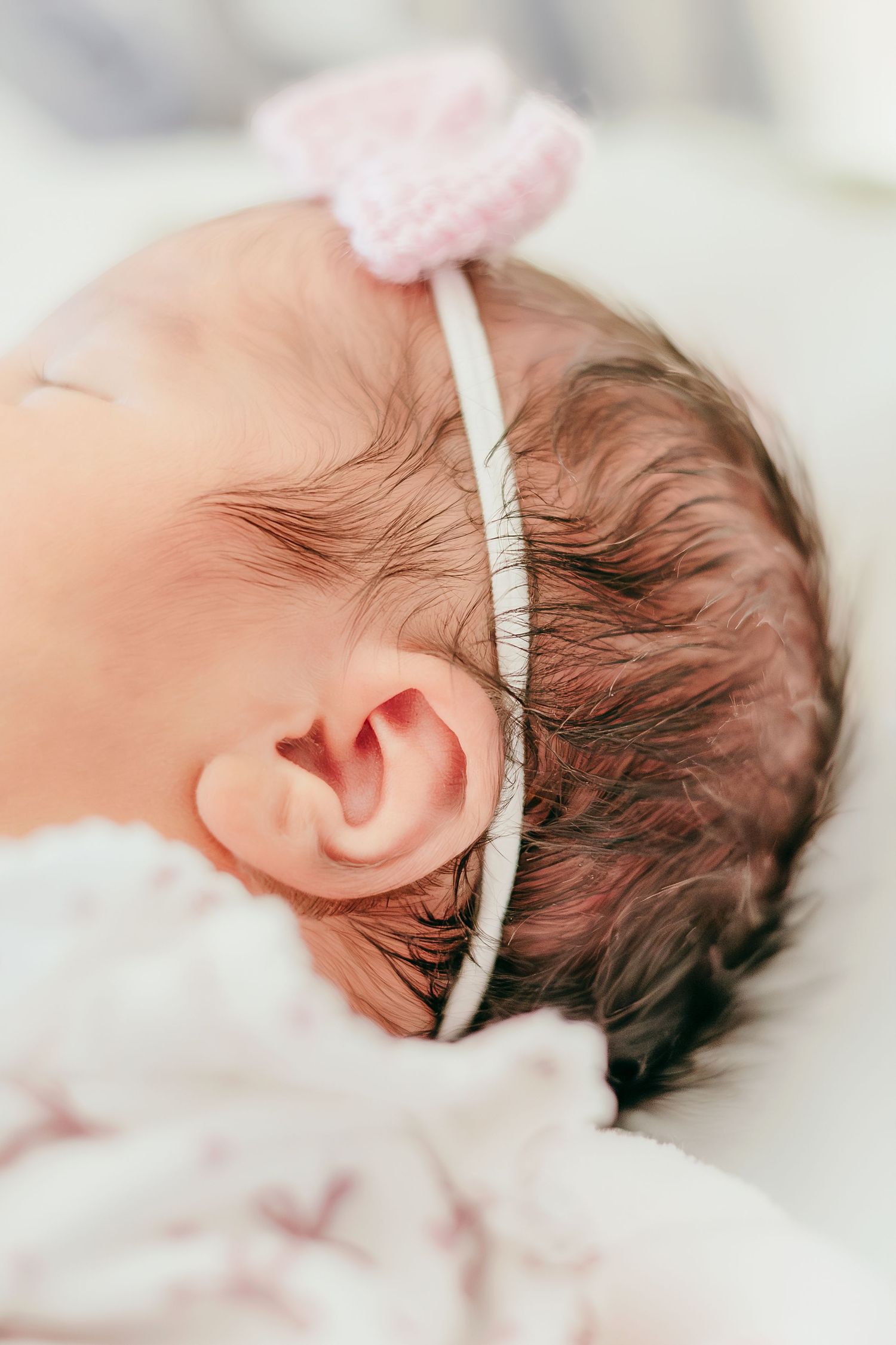 Close-up of a sleeping newborn baby with dark hair wearing a delicate pink crochet bow headband.
