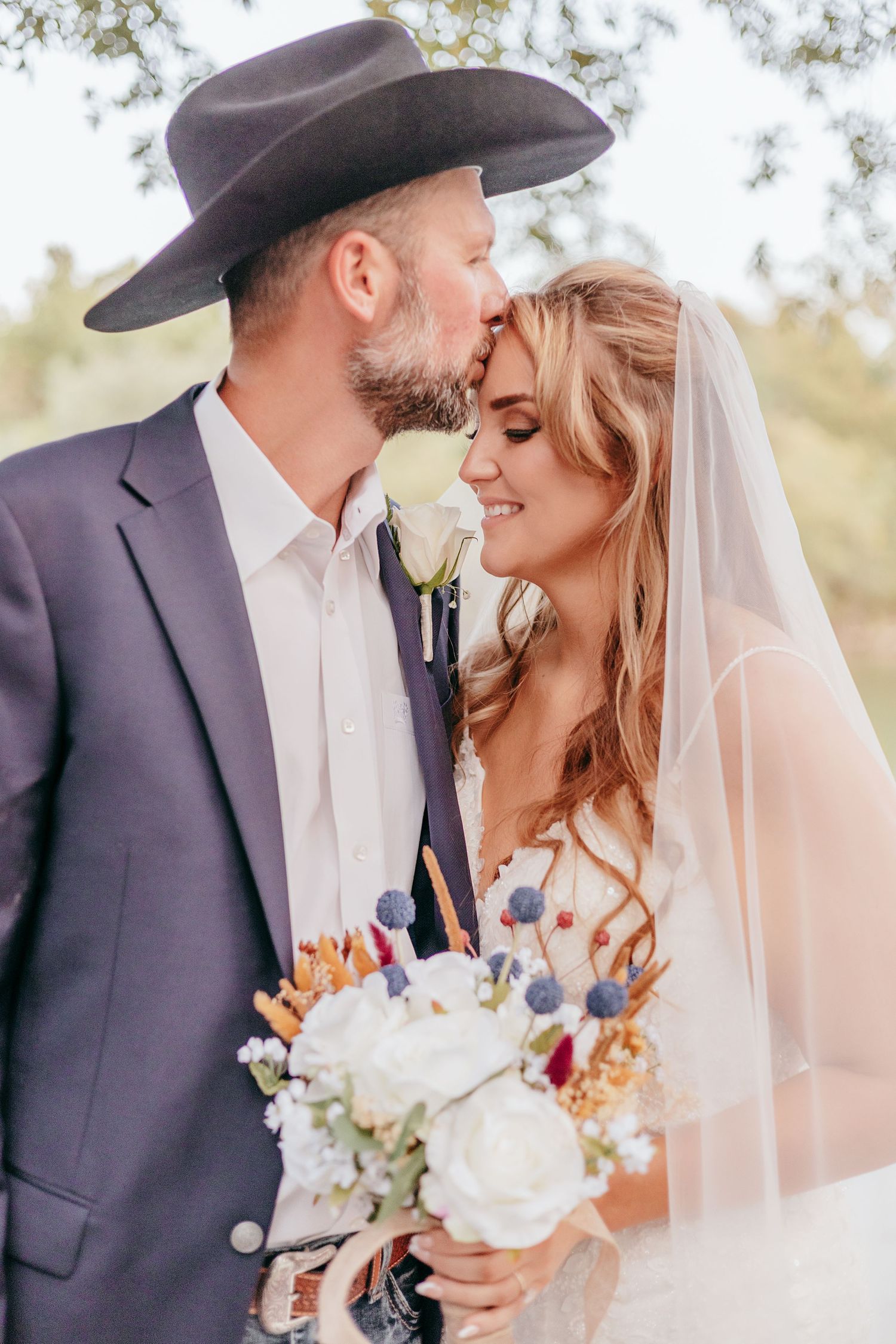 Groom in black cowboy hat kissing smiling bride holding white and blue bouquet.