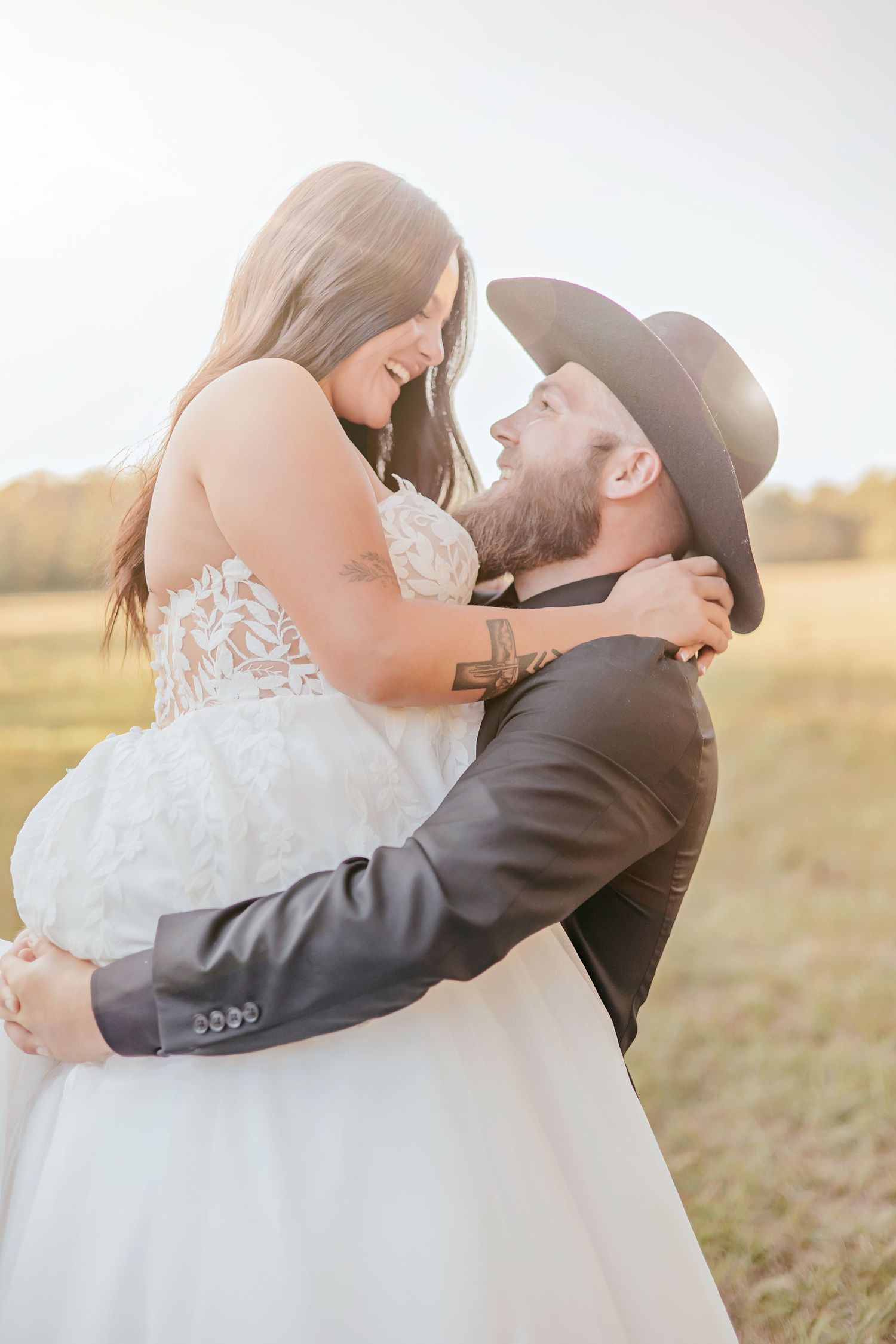 Groom in black cowboy hat lifting smiling bride in lace wedding gown in golden sunlit field.