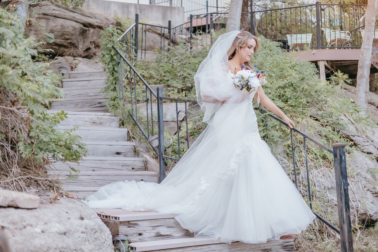Elegant bride in fitted lace wedding gown with long veil descending rustic wooden staircase outdoors.