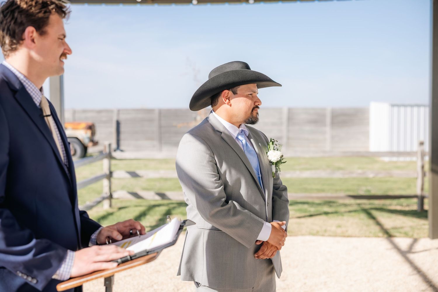 Groom in gray suit and black cowboy hat listens attentively during outdoor wedding ceremony.