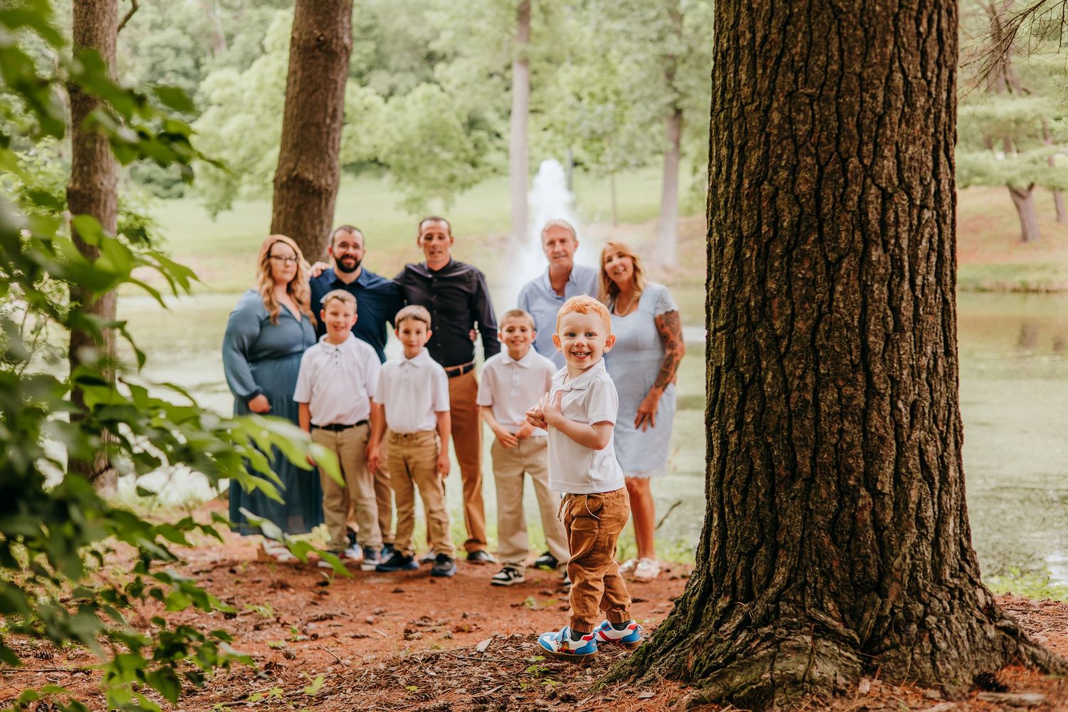 Multi-generational family portrait outdoors in wooded park setting with fountain, featuring grandparents, parents, and young children.