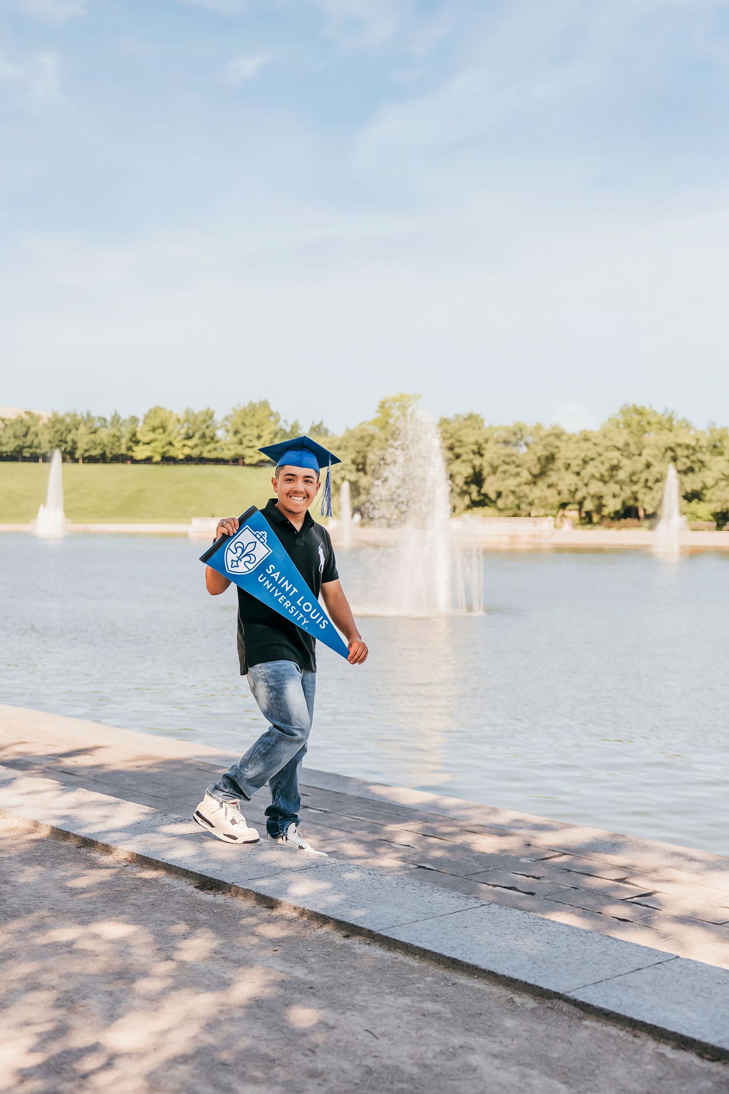 Smiling graduate in blue cap holding a university pennant flag by a fountain on a sunny day.