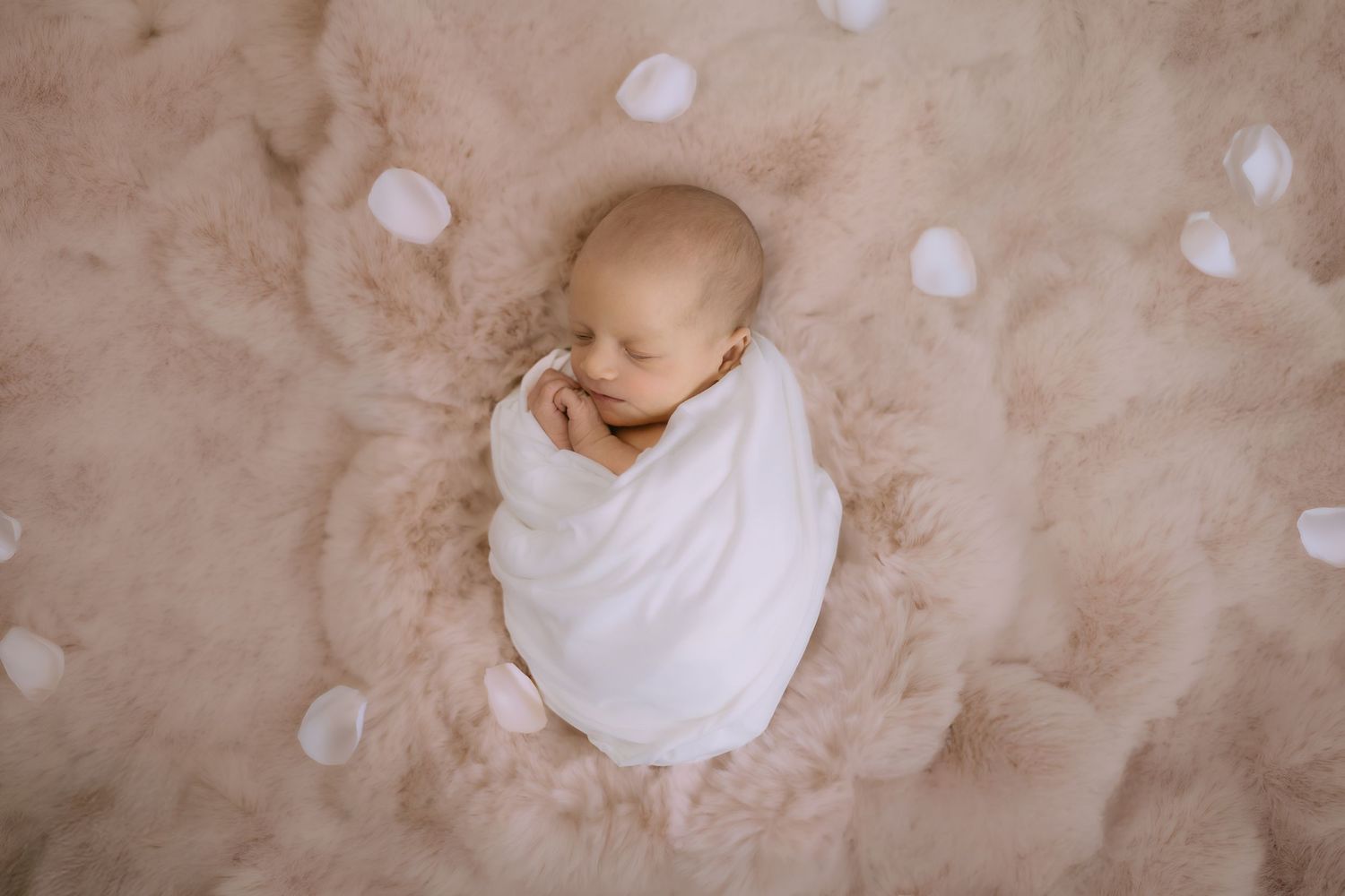 Newborn baby swaddled in white wrap lying on a fluffy beige fur blanket.