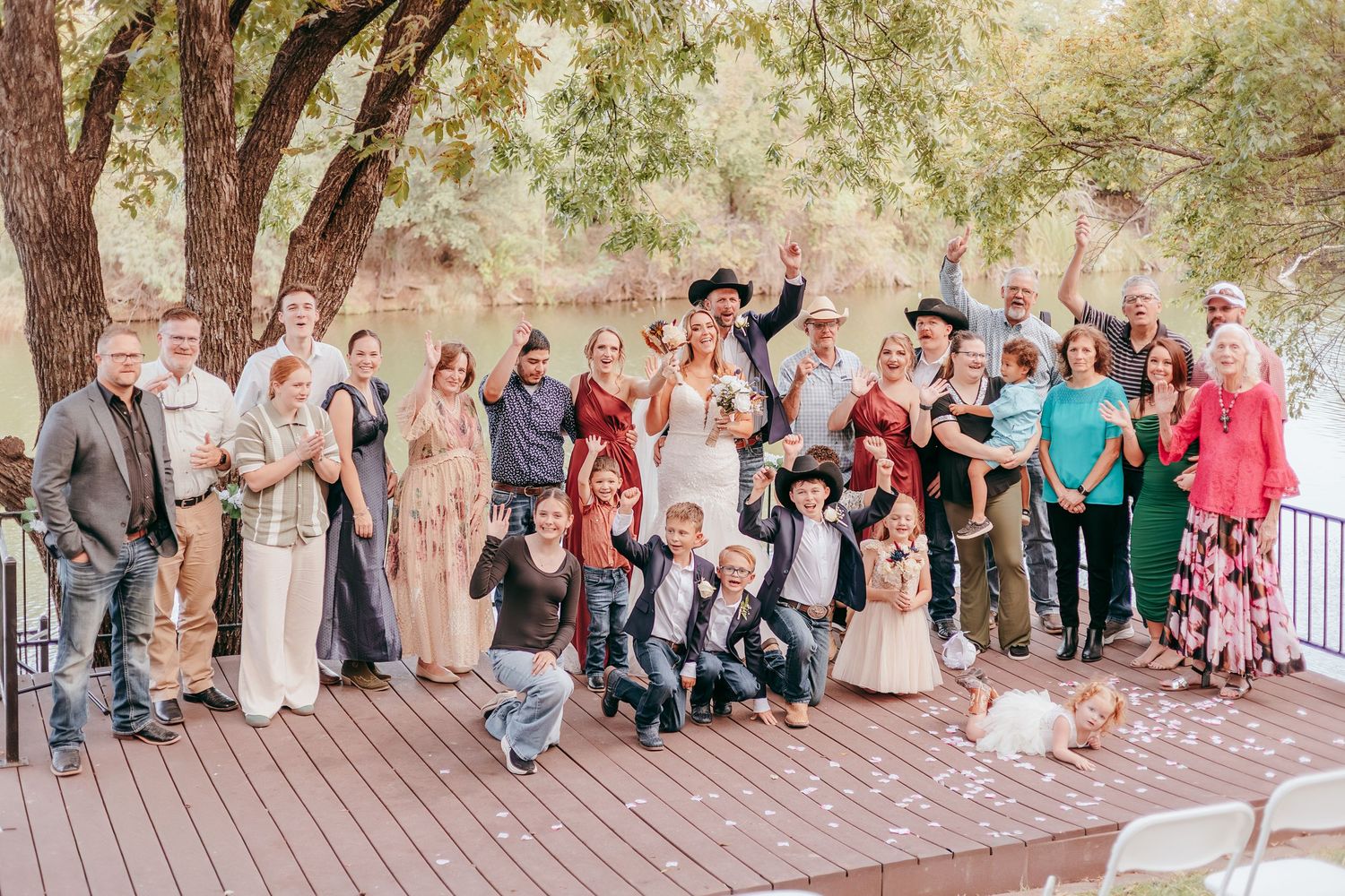 Large wedding party group photo on wooden deck by a tree-lined river.