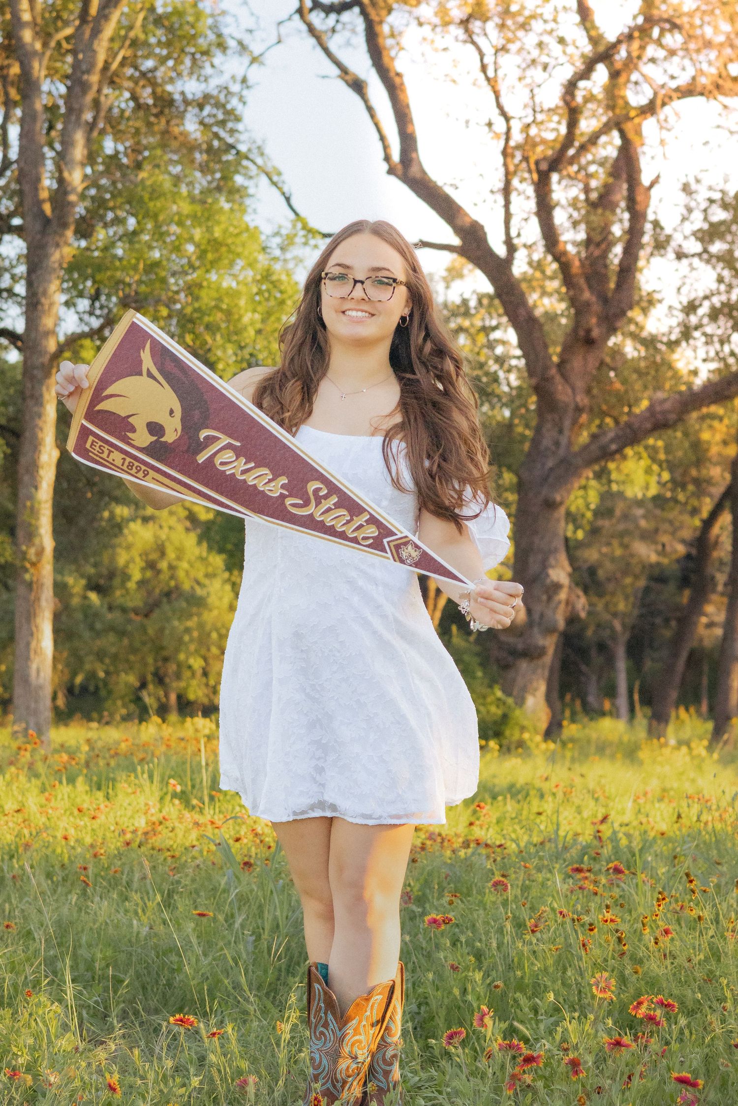 Young woman in white dress and cowboy boots holding a Texas State University pennant in a sunny field.