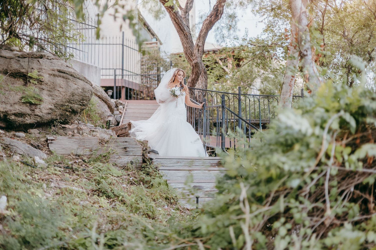 Elegant bride in lace ball gown wedding dress standing on outdoor steps surrounded by lush green foliage.