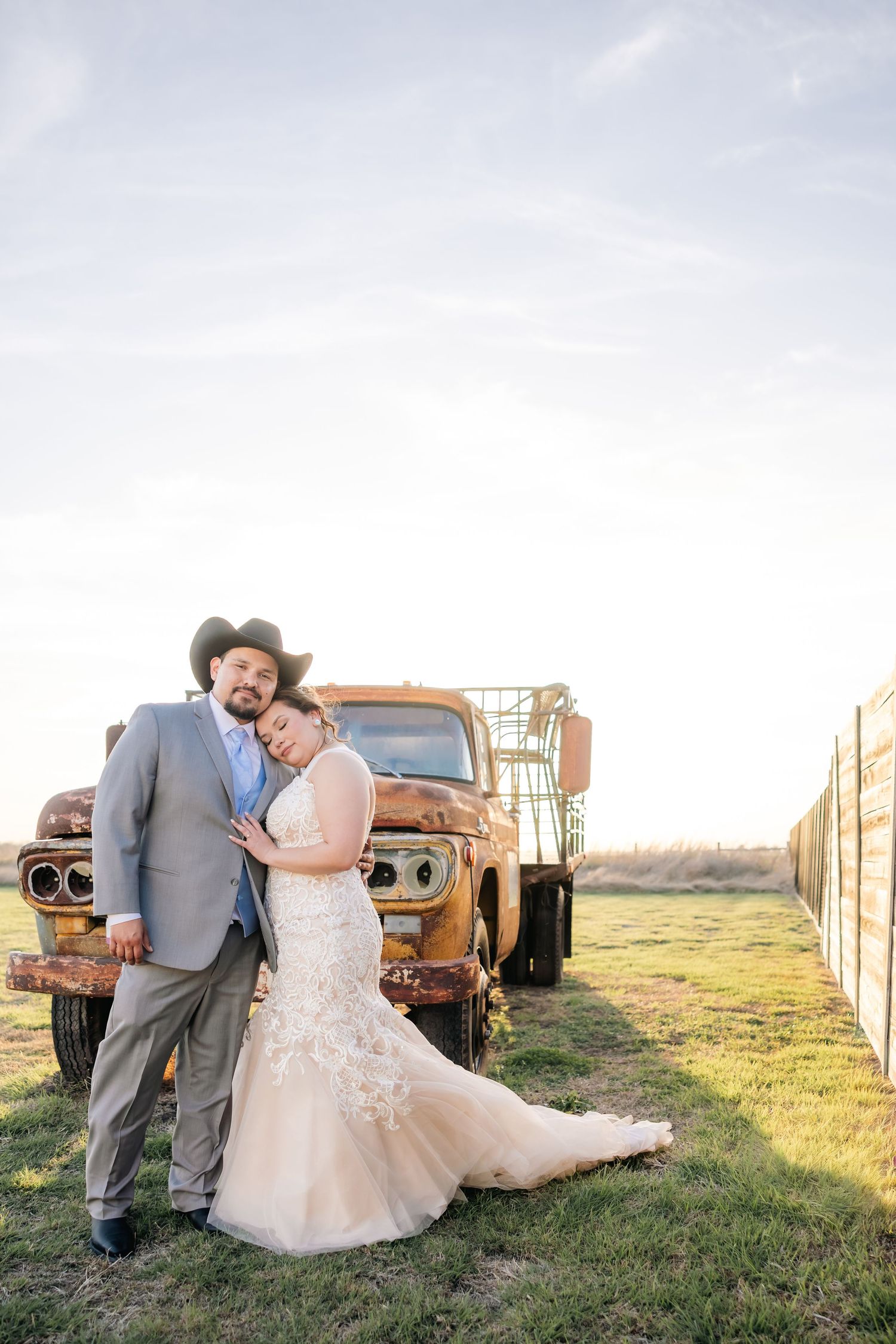Groom in cowboy hat embraces bride in lace mermaid gown leaning on a vintage rusty truck at sunset.