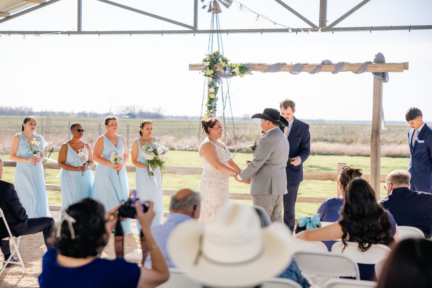 Bride and groom exchange vows under rustic pergola with bridesmaids in light blue dresses.