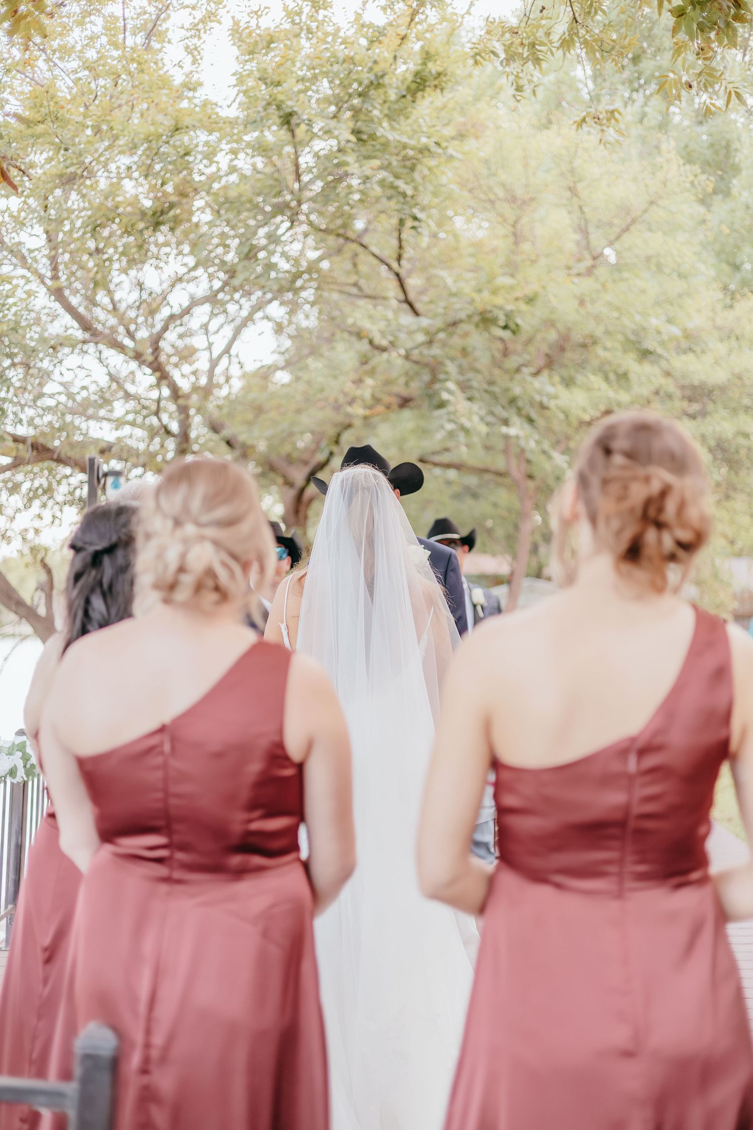 Bride in white gown and veil walks with bridesmaids in dusty rose dresses toward outdoor wedding ceremony.