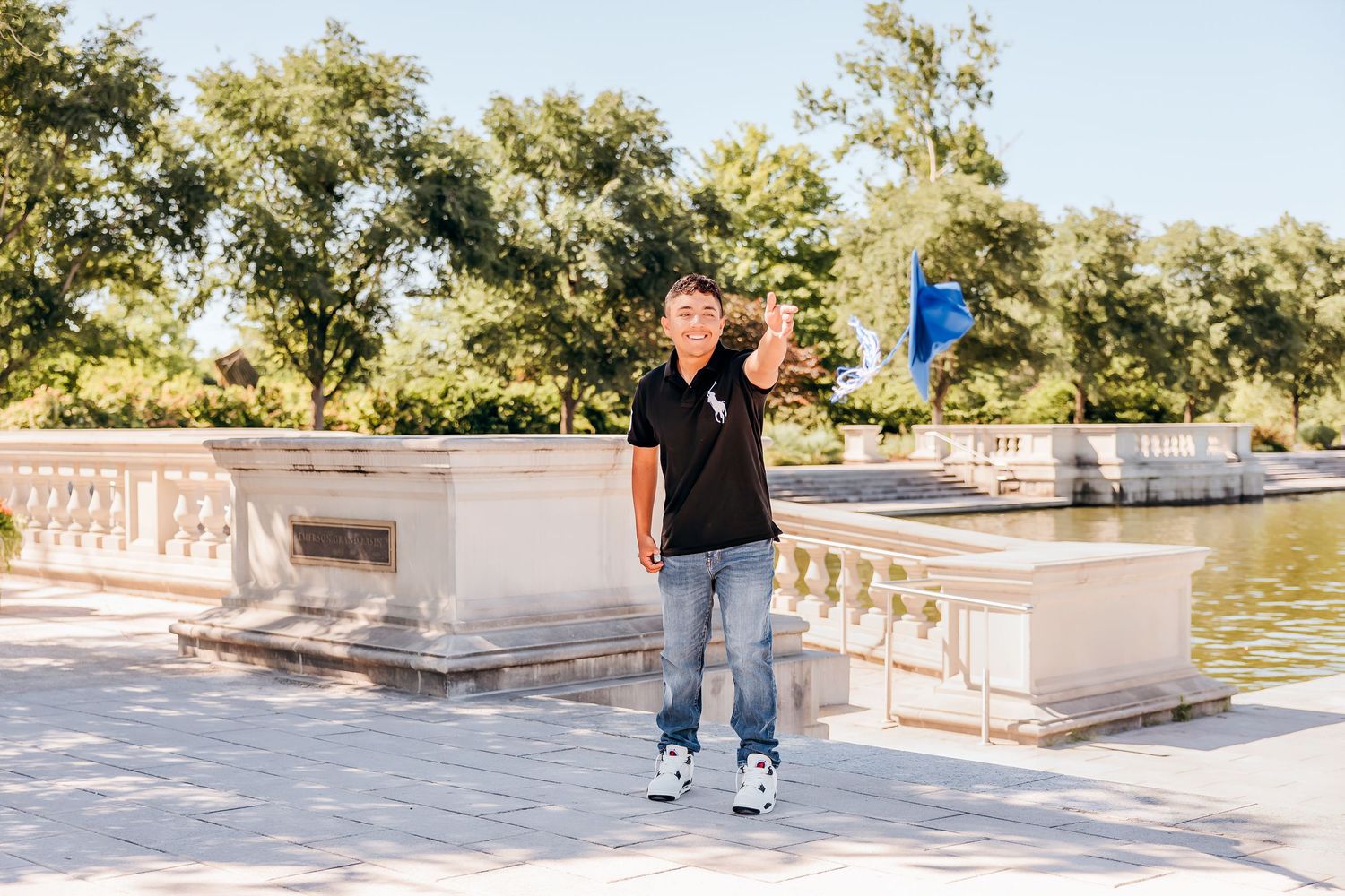 Young man in black polo and jeans poses near ornate white stone fountain in sunny park setting.