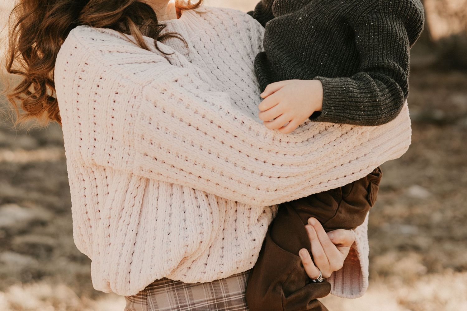 A mother in a pink knit sweater lovingly holds her young child outdoors in warm light.