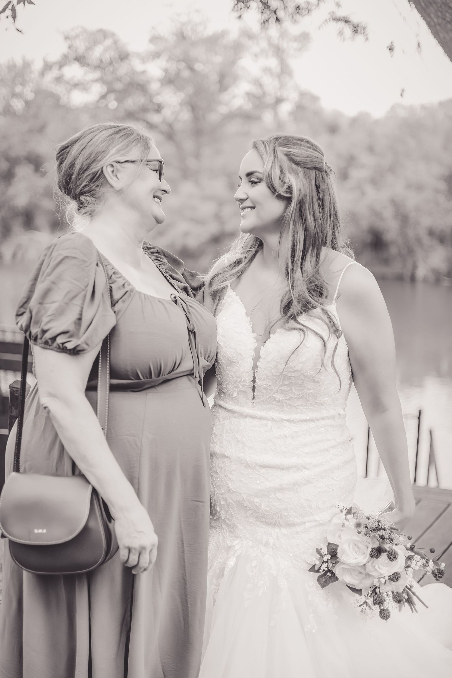 Black and white photo of bride and older woman smiling at each other by a river.