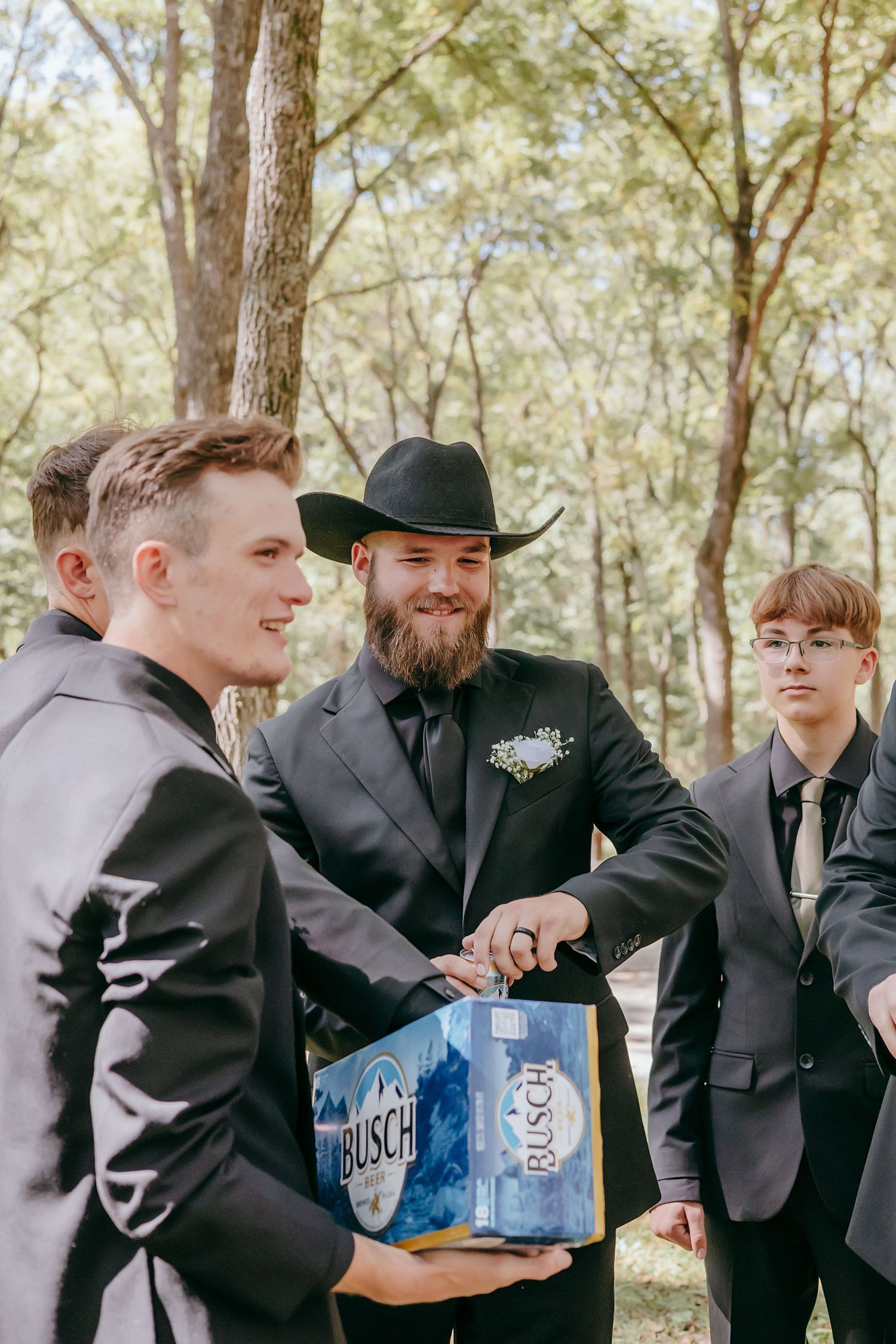 Bearded groom in black suit and cowboy hat sharing Busch beer case with groomsmen in wooded outdoor setting.