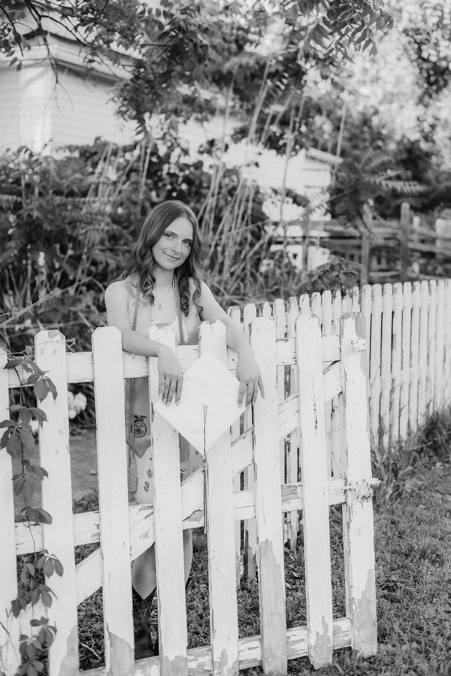 Young woman in white dress leaning on a white picket fence surrounded by lush garden greenery, black and white photo.