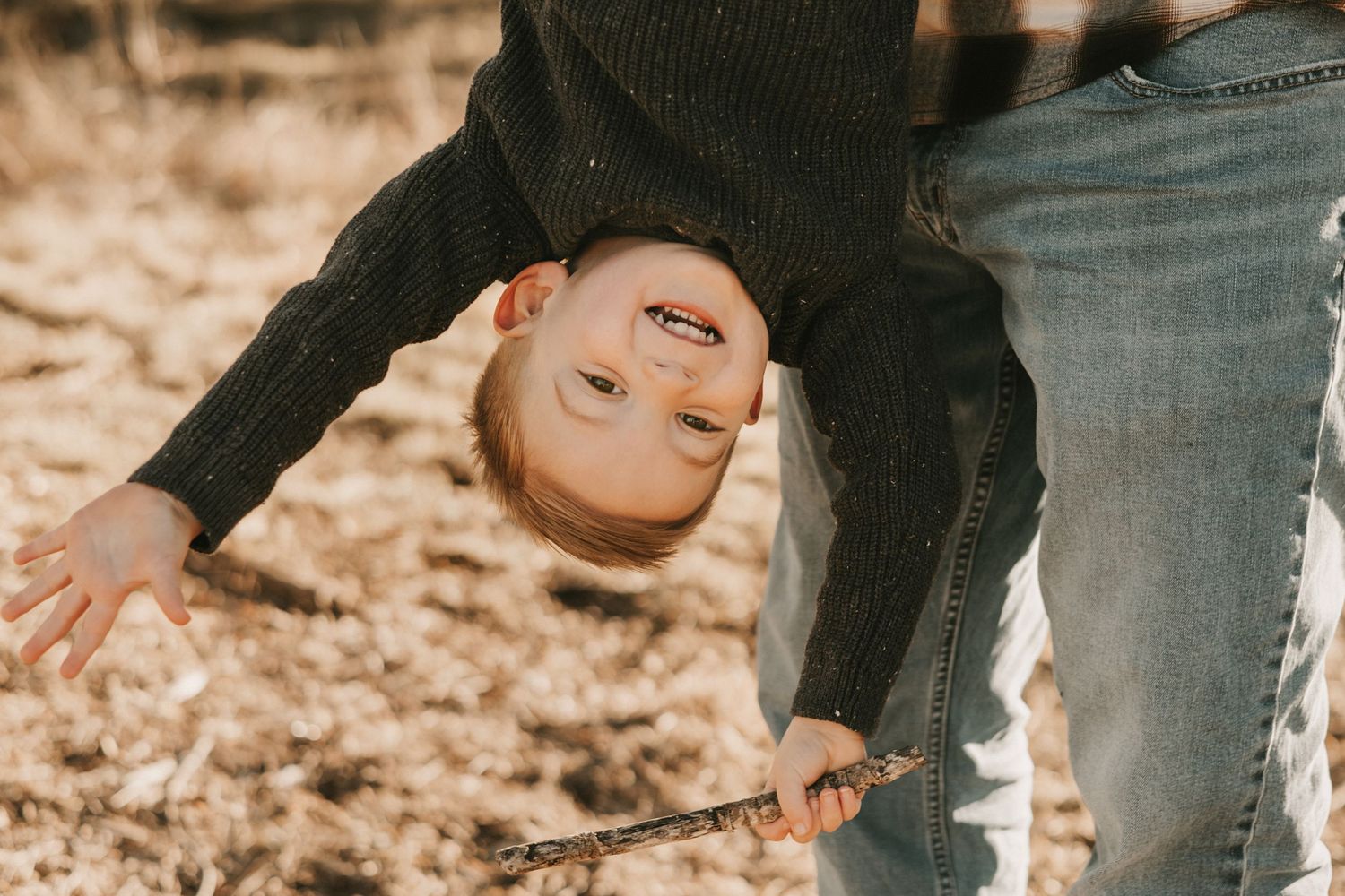 A young child hangs upside down playfully outdoors, smiling while holding a stick in autumn surroundings.