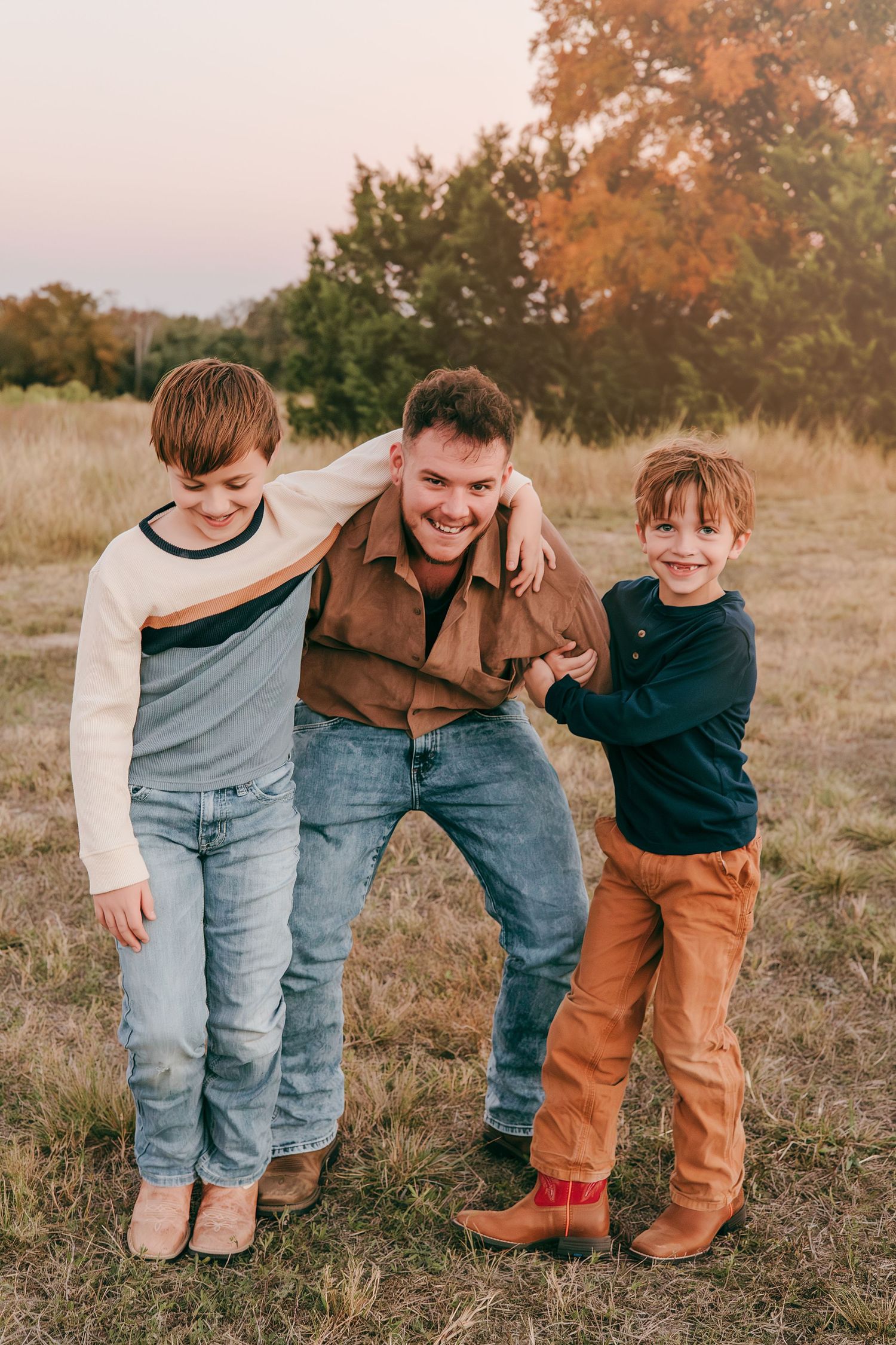 A smiling man bends down as two young boys wrap their arms around him in an autumn outdoor family portrait.