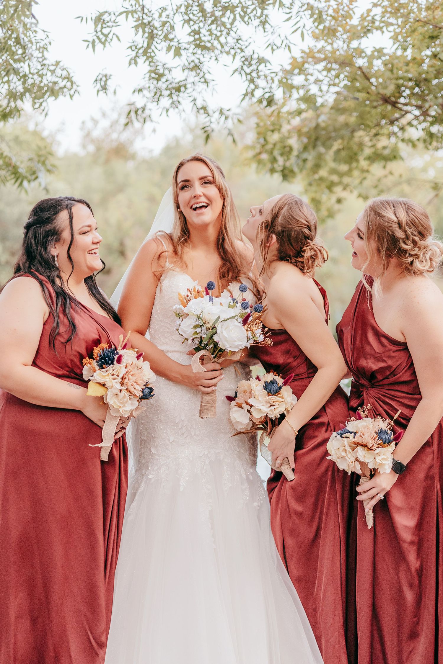 Laughing bride in white gown with three bridesmaids in burgundy dresses outdoors.