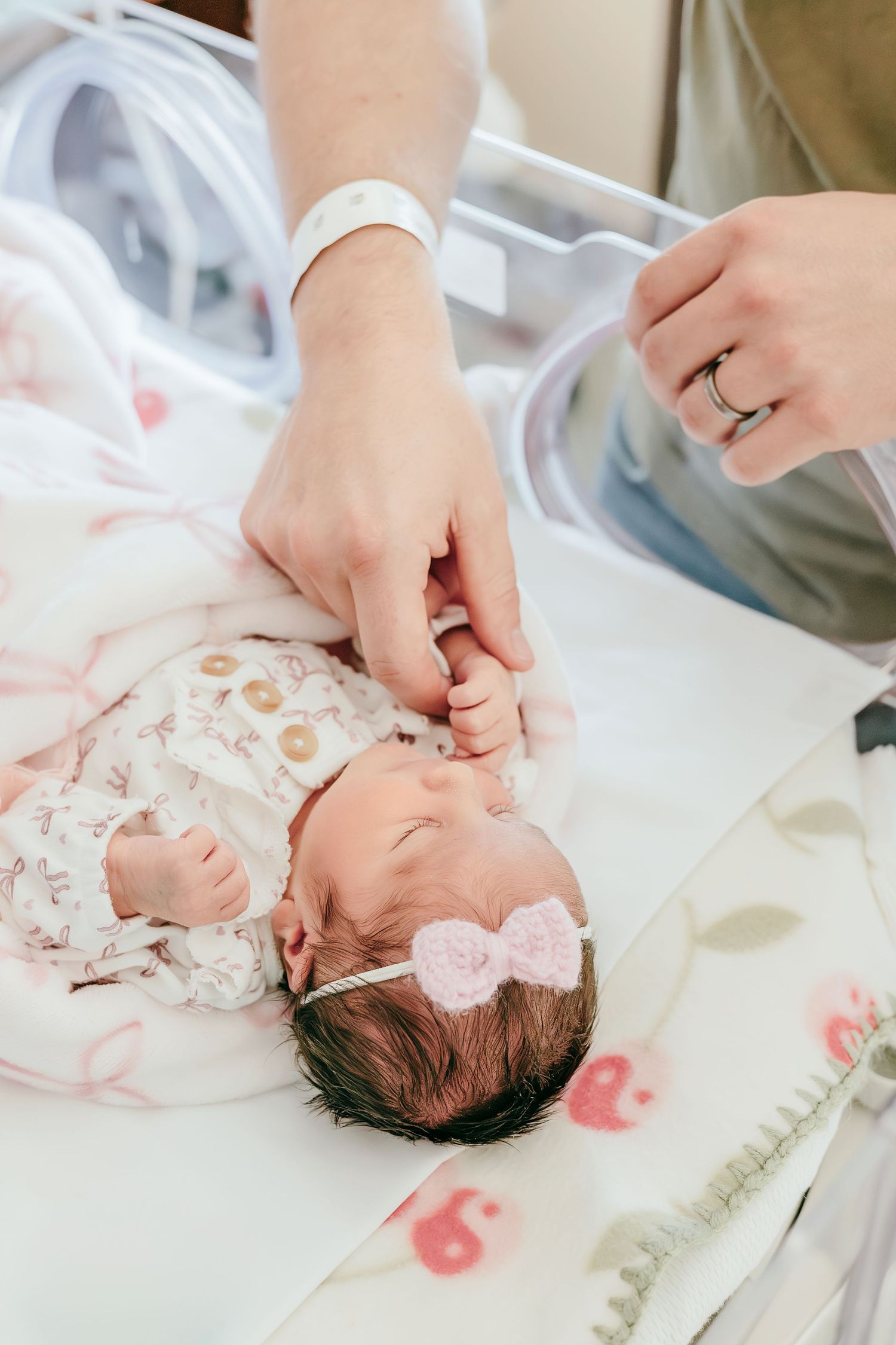 Newborn baby in floral onesie and pink bow headband gently gripping a parent's finger in hospital.